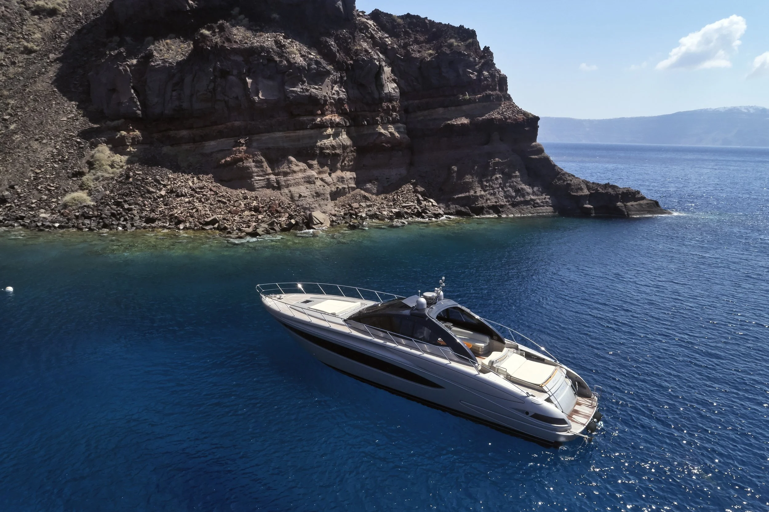 A yacht floating on blue water near a rocky coastline with cliffs under a partly cloudy sky.