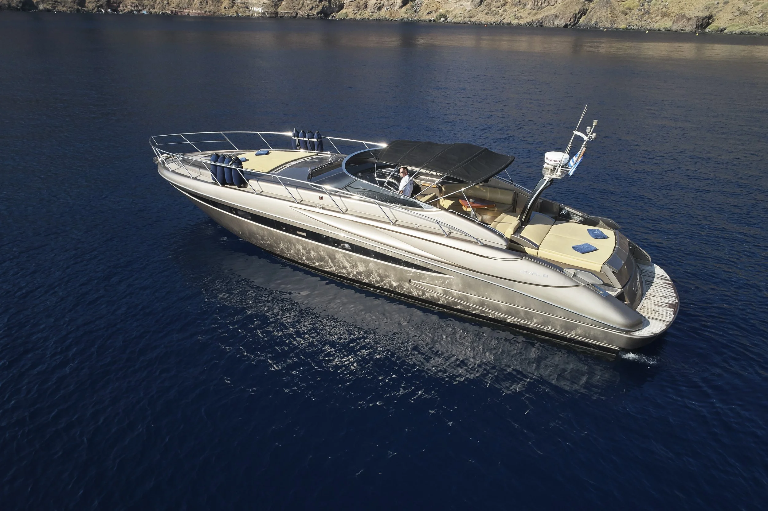 A large luxurious yacht sailing on a calm body of water with a rocky shoreline in the background. The yacht has a black canopy over the center and beige seating areas. There's a person at the helm.