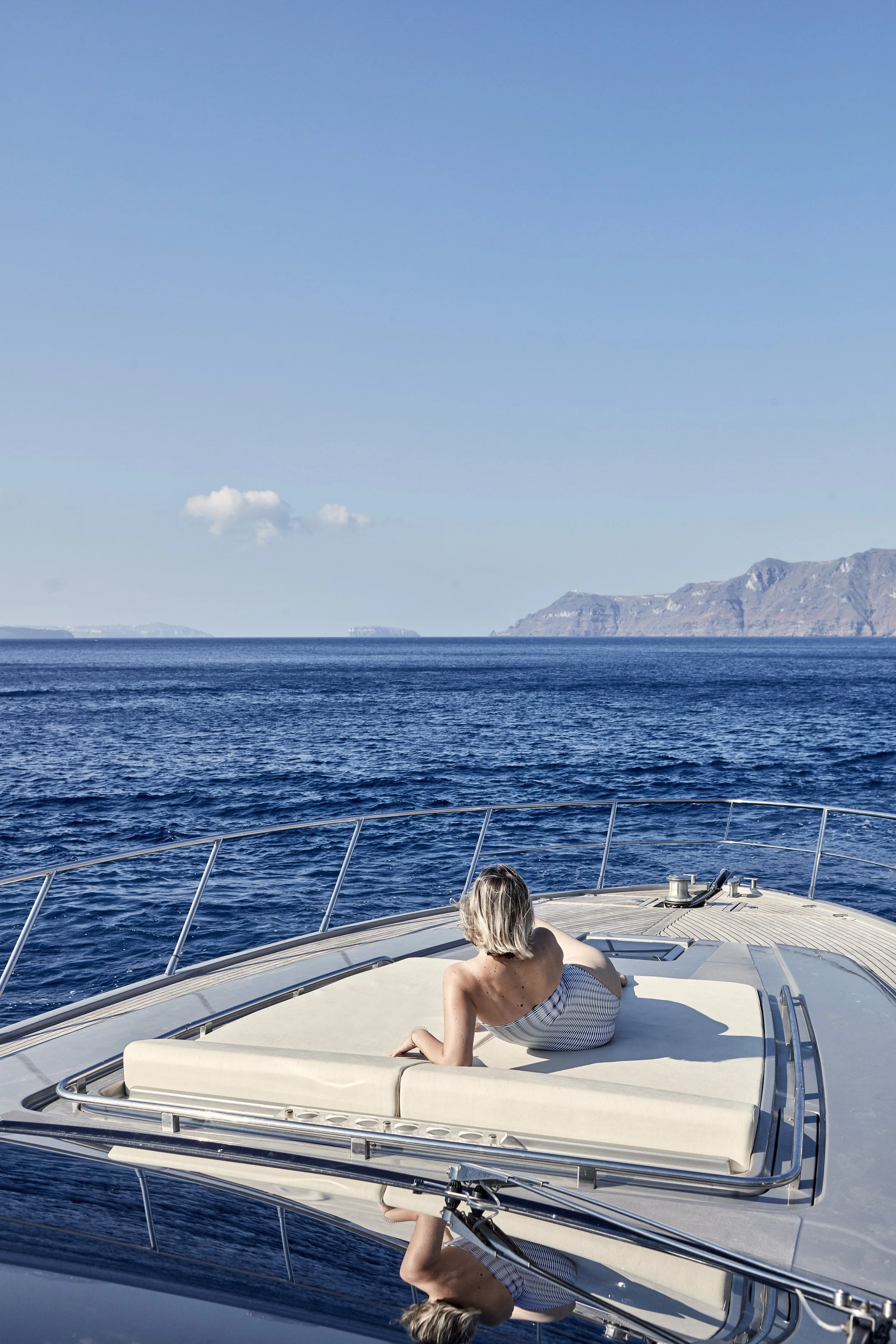 A woman with blonde hair, wearing a striped white and gray swimsuit, is lying on her side on a boat deck, facing the ocean, with mountains visible in the distance under a clear blue sky.