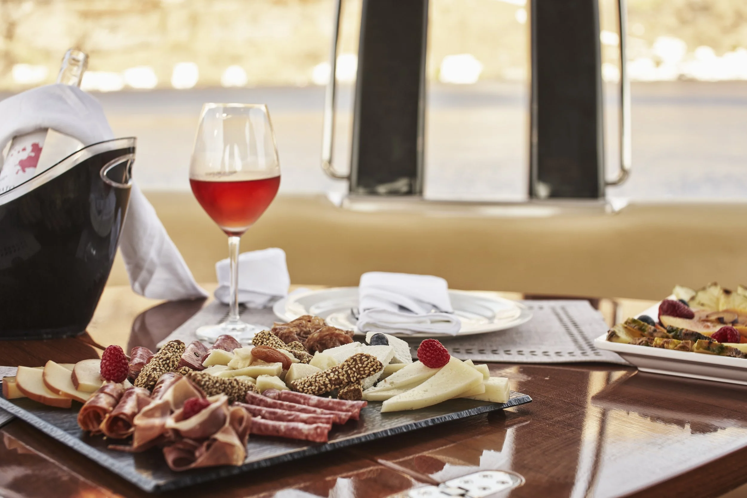 A table set with a cheese and charcuterie platter, a glass of red wine, a napkin, and a white plate with food, with a window showing an outdoor scene in the background.