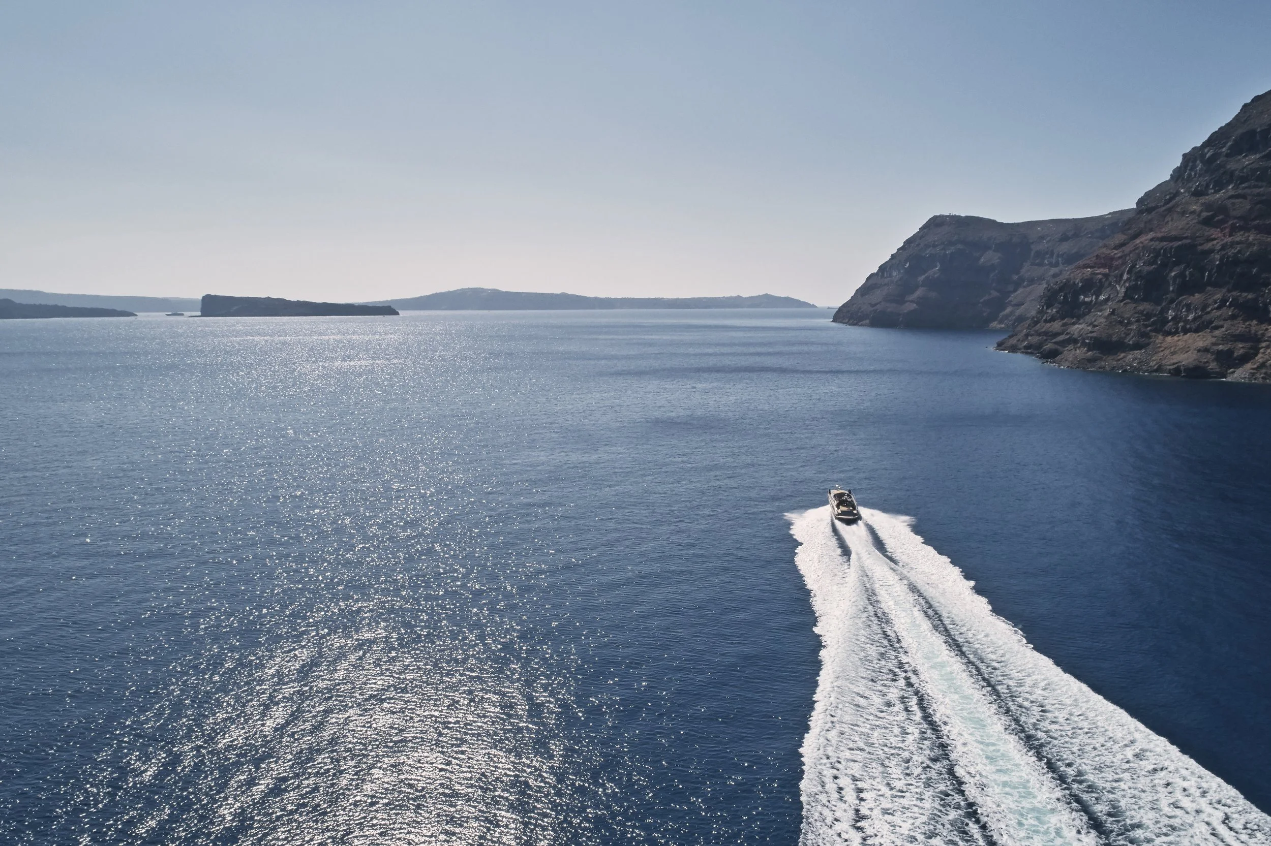 A boat speeds through a large body of water, leaving a white wake behind, with cliffs and landforms on the horizon under a clear sky.