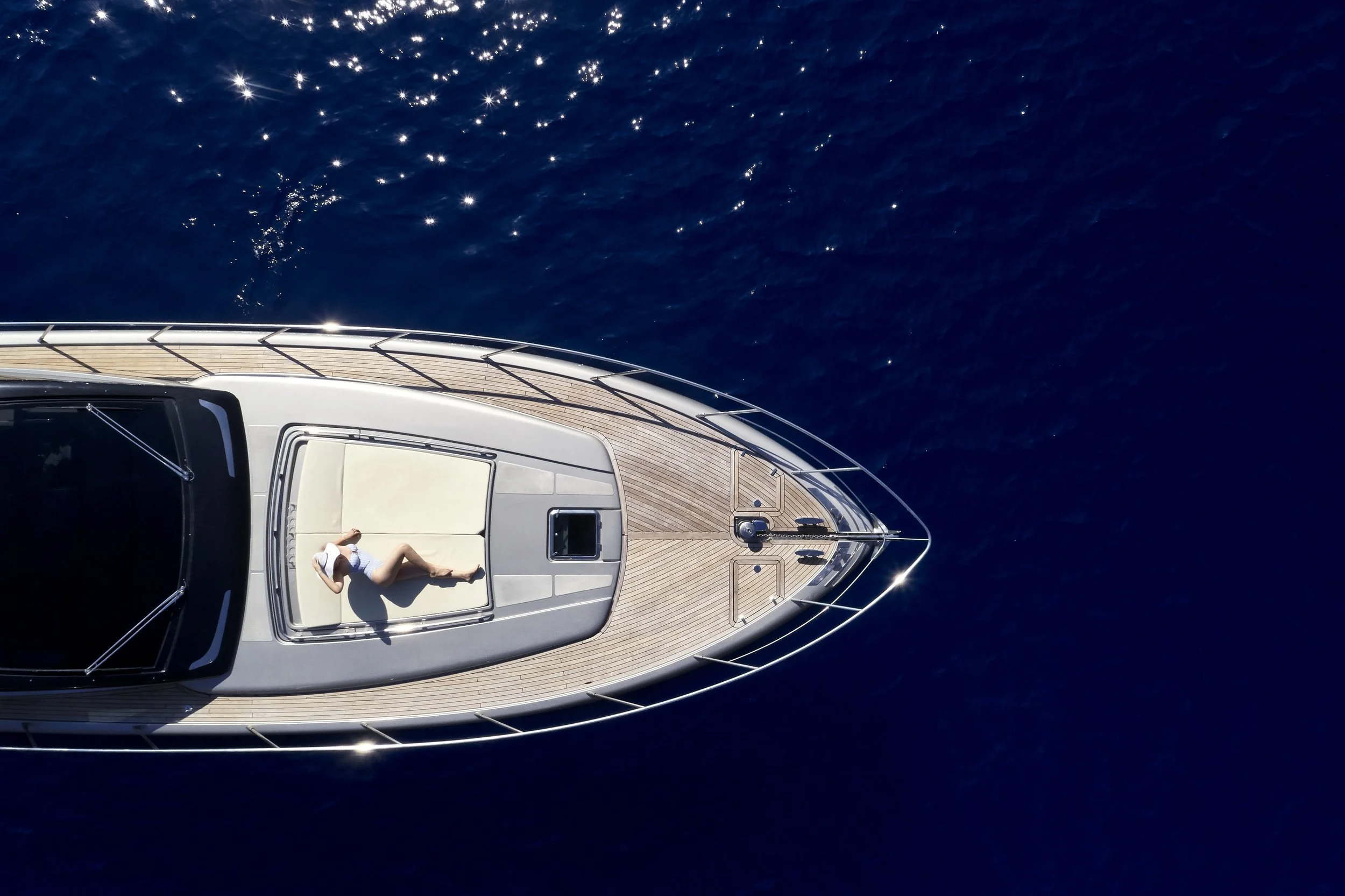 Top-down view of a person lying on a sunbathing pad on the deck of a yacht in the open water.