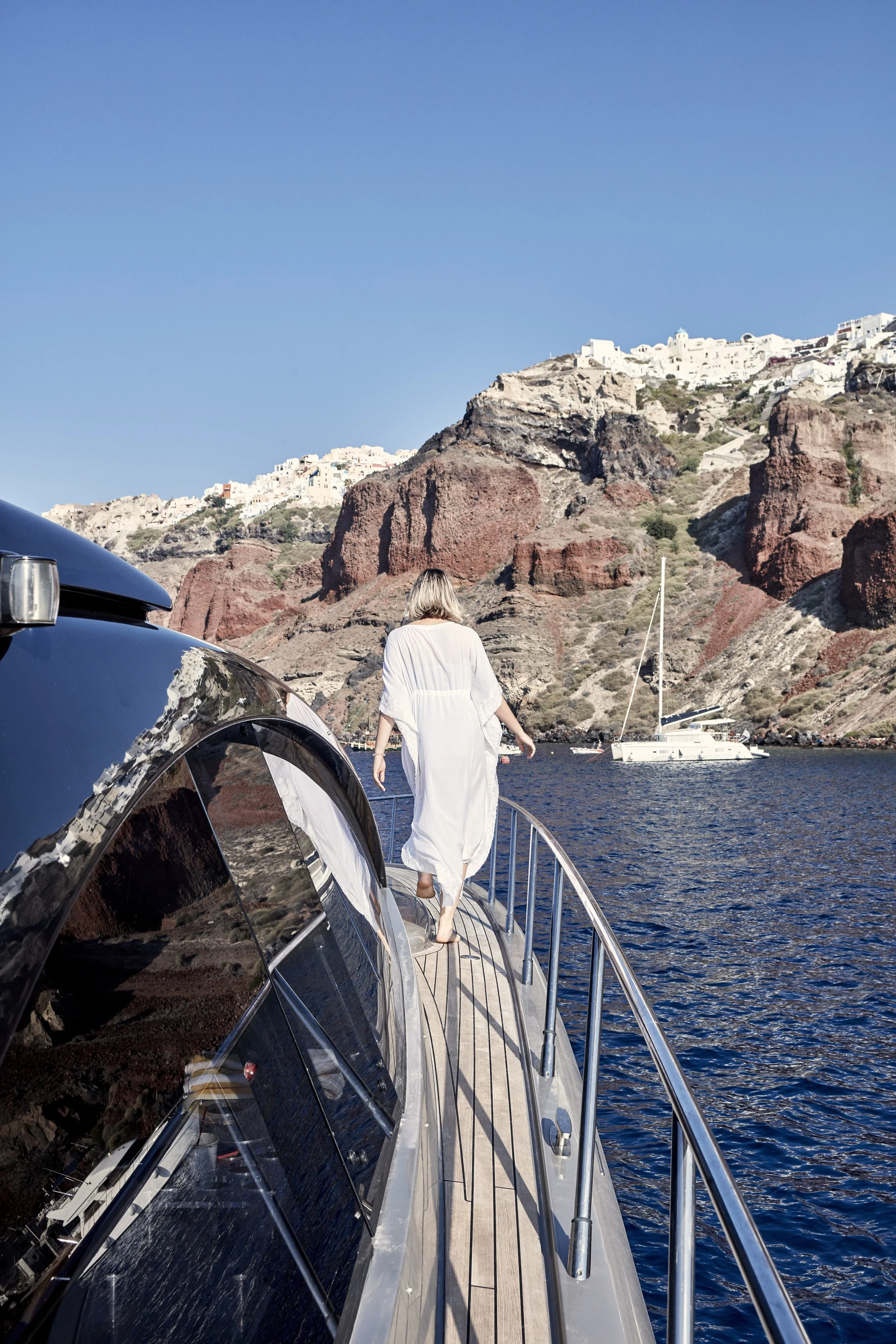 Woman walking on a yacht with a coastal landscape behind her, featuring rocks and white buildings.