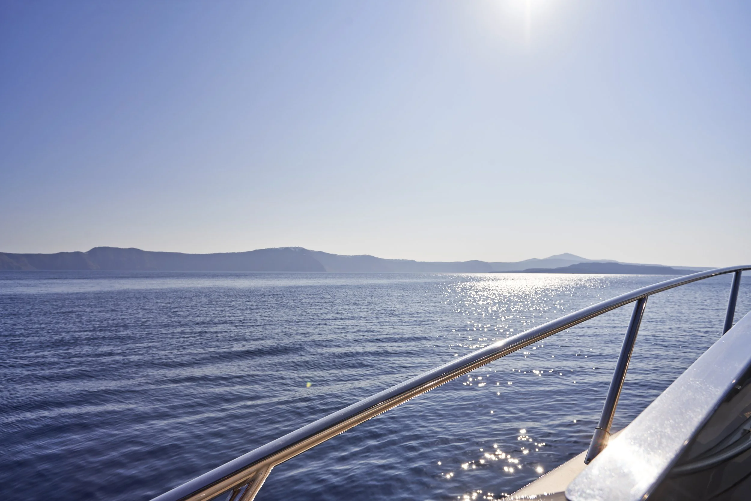View of the ocean from a boat, with distant land and mountains on the horizon, and sunlight reflecting off the water.