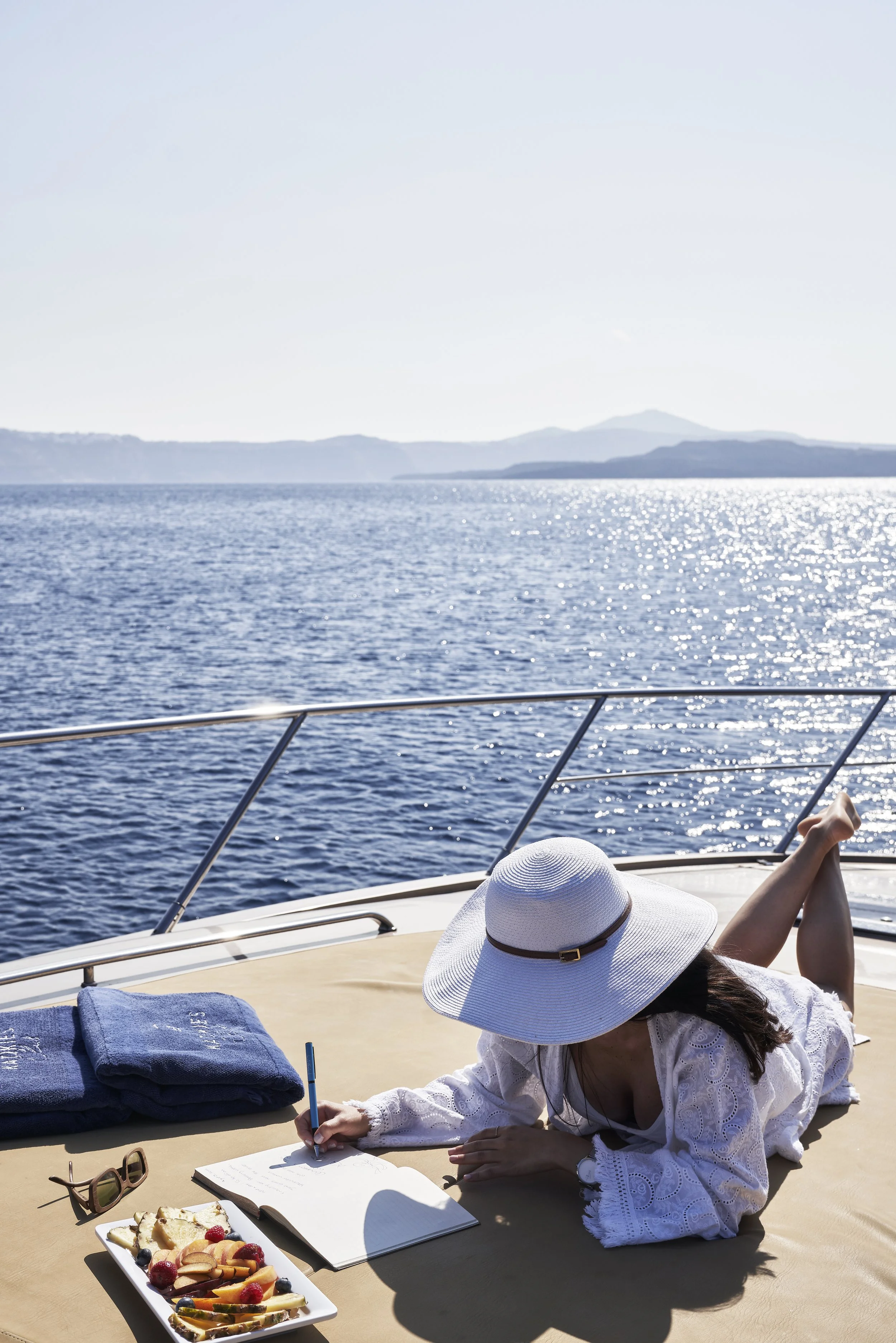 A woman lying on a yacht, writing in a book with a pen. She is wearing a large sun hat, sunglasses, and a white dress. There is a plate of assorted fruit and snacks, towels, and her sunglasses nearby. The yacht is on a calm, sparkling blue sea with d