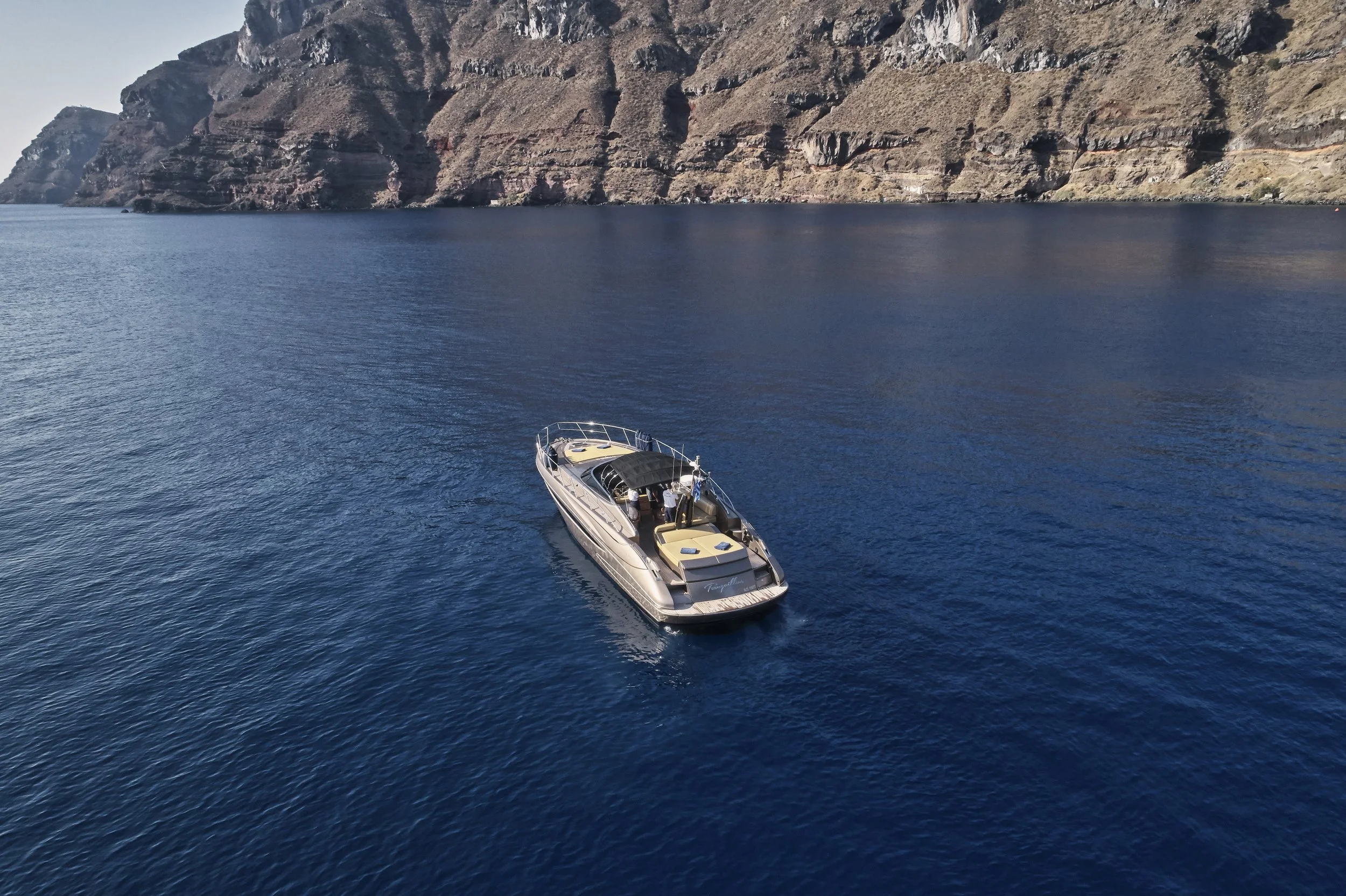 A yacht sailing on a calm, deep blue body of water with rocky, mountainous shoreline in the background.