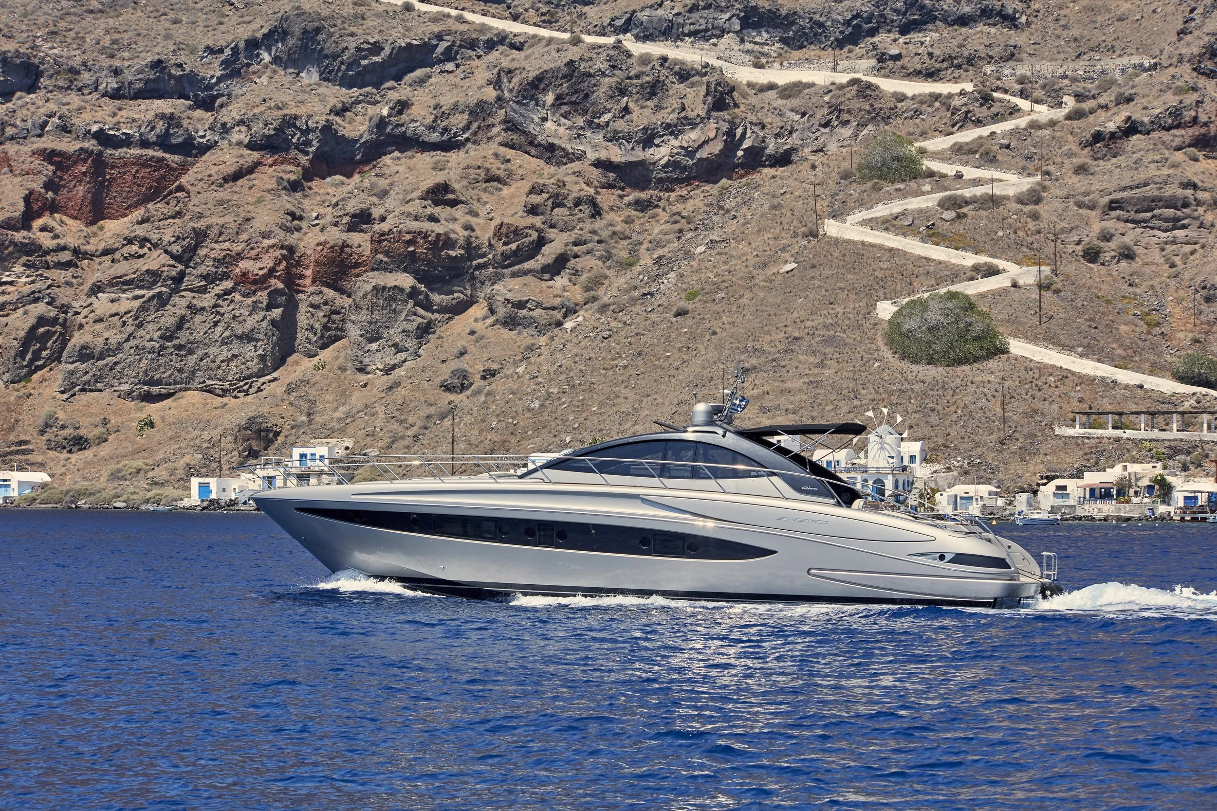 A sleek, modern yacht cruising on blue water, with dry, mountainous terrain and white buildings along the shoreline in the background.