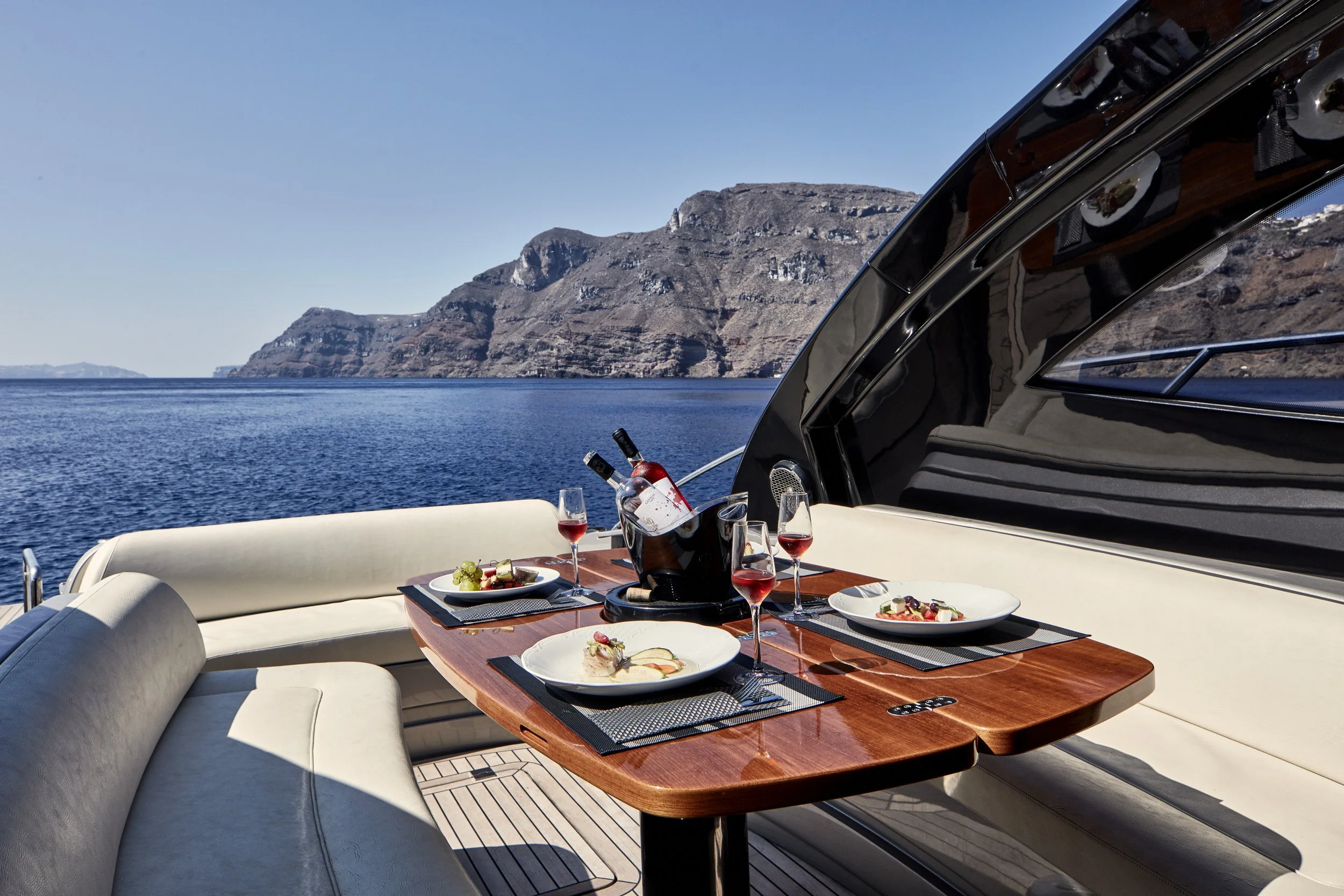 A yacht dining area with a wooden table set with plates of food, glasses of red wine, and a bucket with wine bottles, overlooking the water and rocky cliffs in the background.