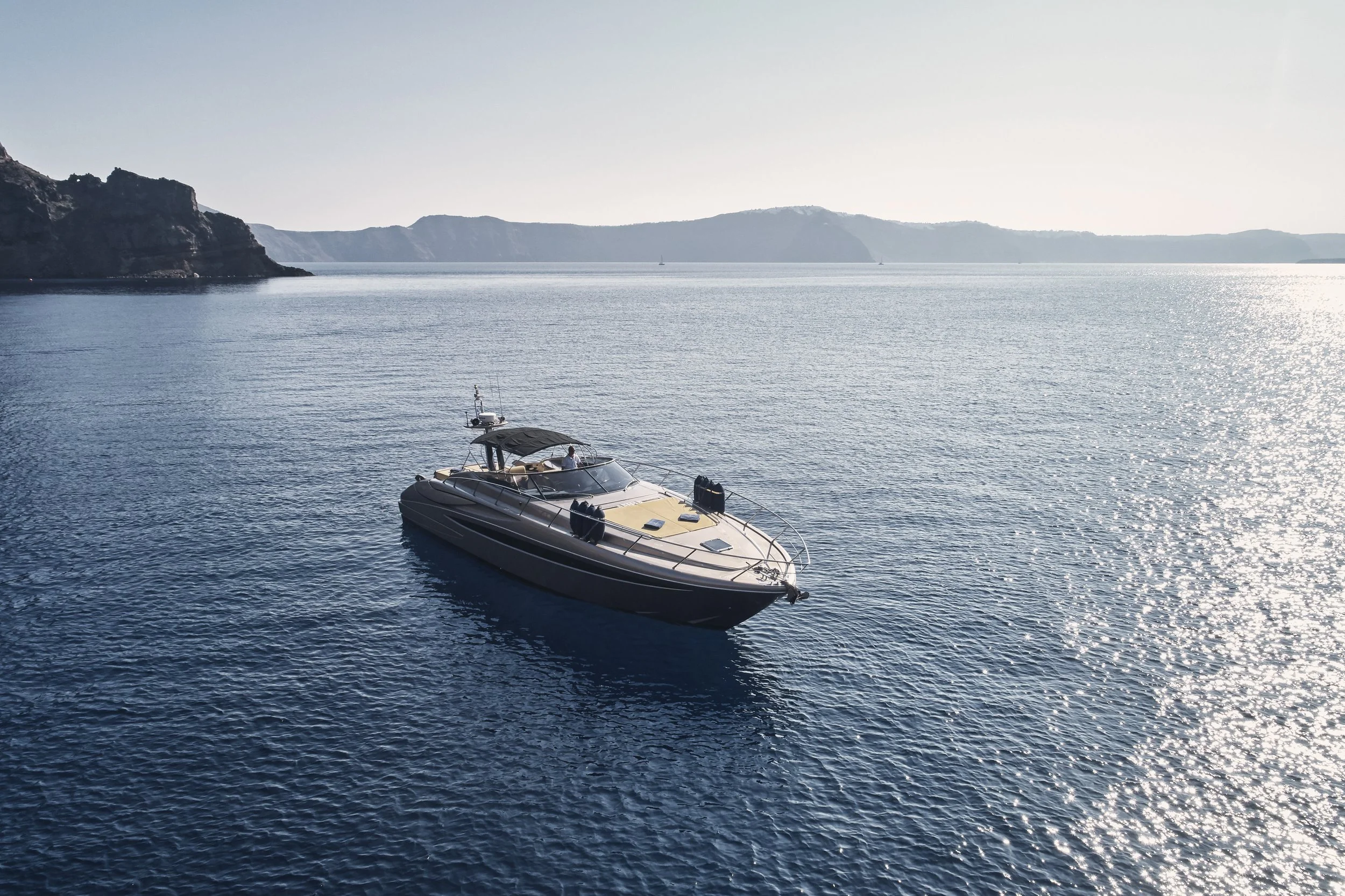 A luxury motor yacht floating on calm ocean water with distant shoreline and cliffs in background, sunlight reflecting off the water.
