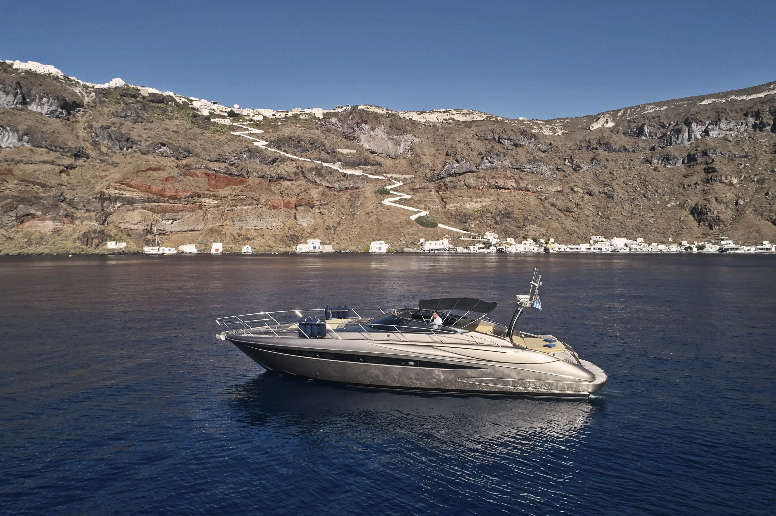 A luxury motorboat cruising on calm deep blue waters with a coastal hillside and white buildings in the background.
