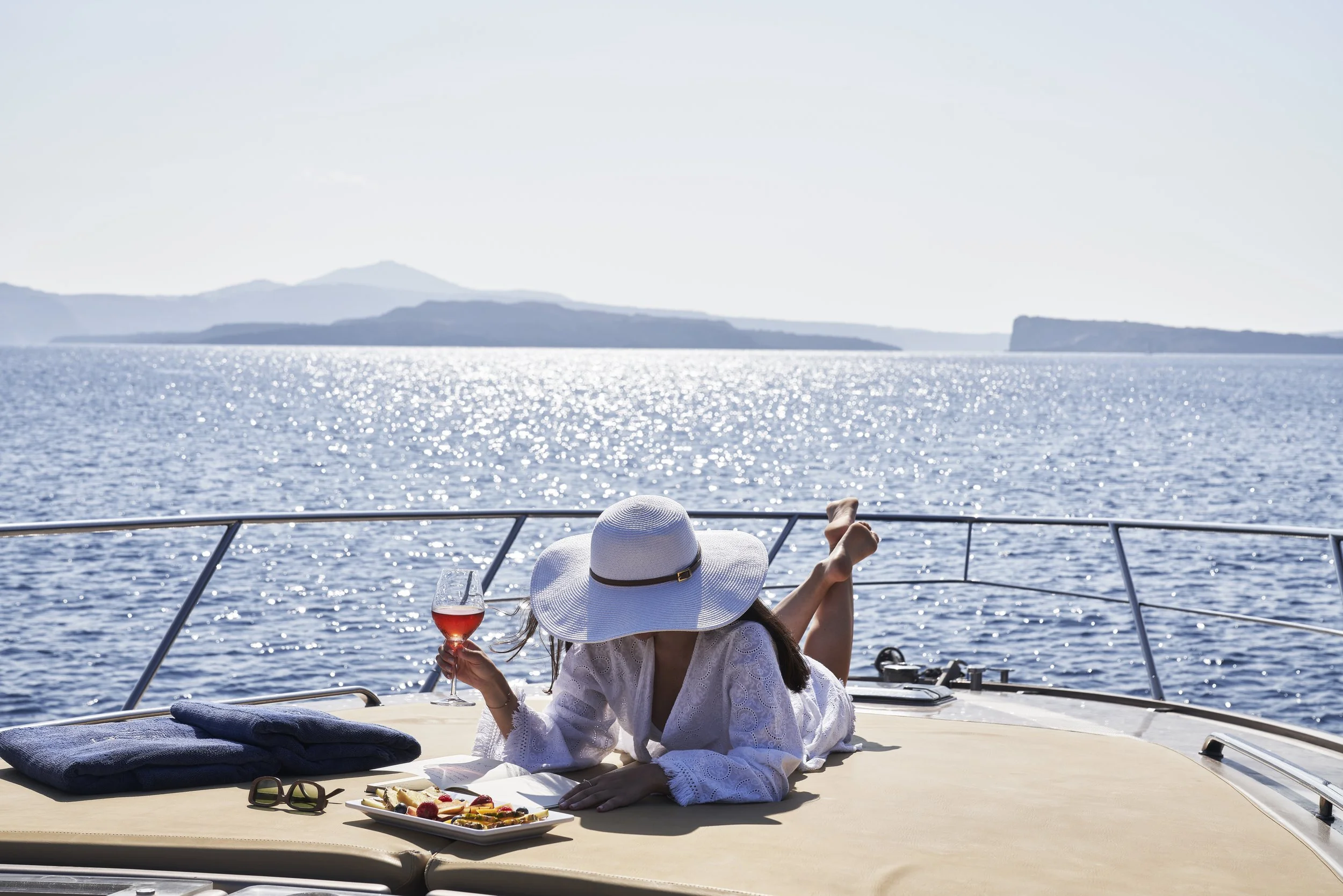 Woman lying on a yacht deck holding a glass of rosé wine, wearing a large sun hat and white cover-up, with a plate of snacks, sunglasses, and a towel nearby, overlooking the ocean with distant islands.