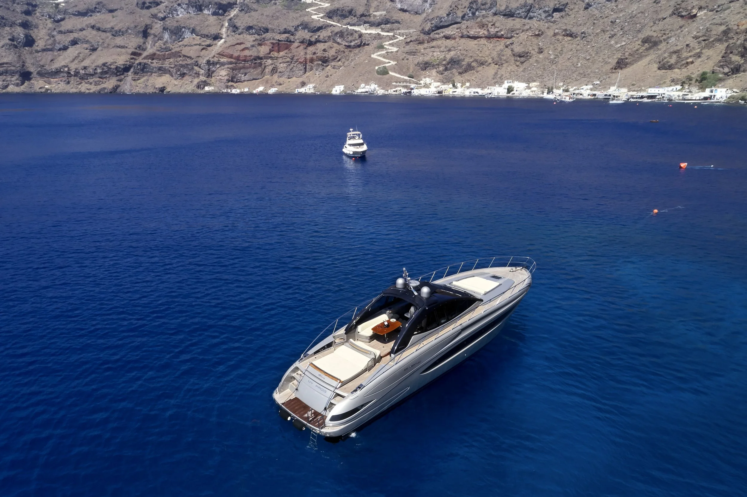 A luxurious yacht floating on deep blue water near a rocky coastline with a small town or village in the background.
