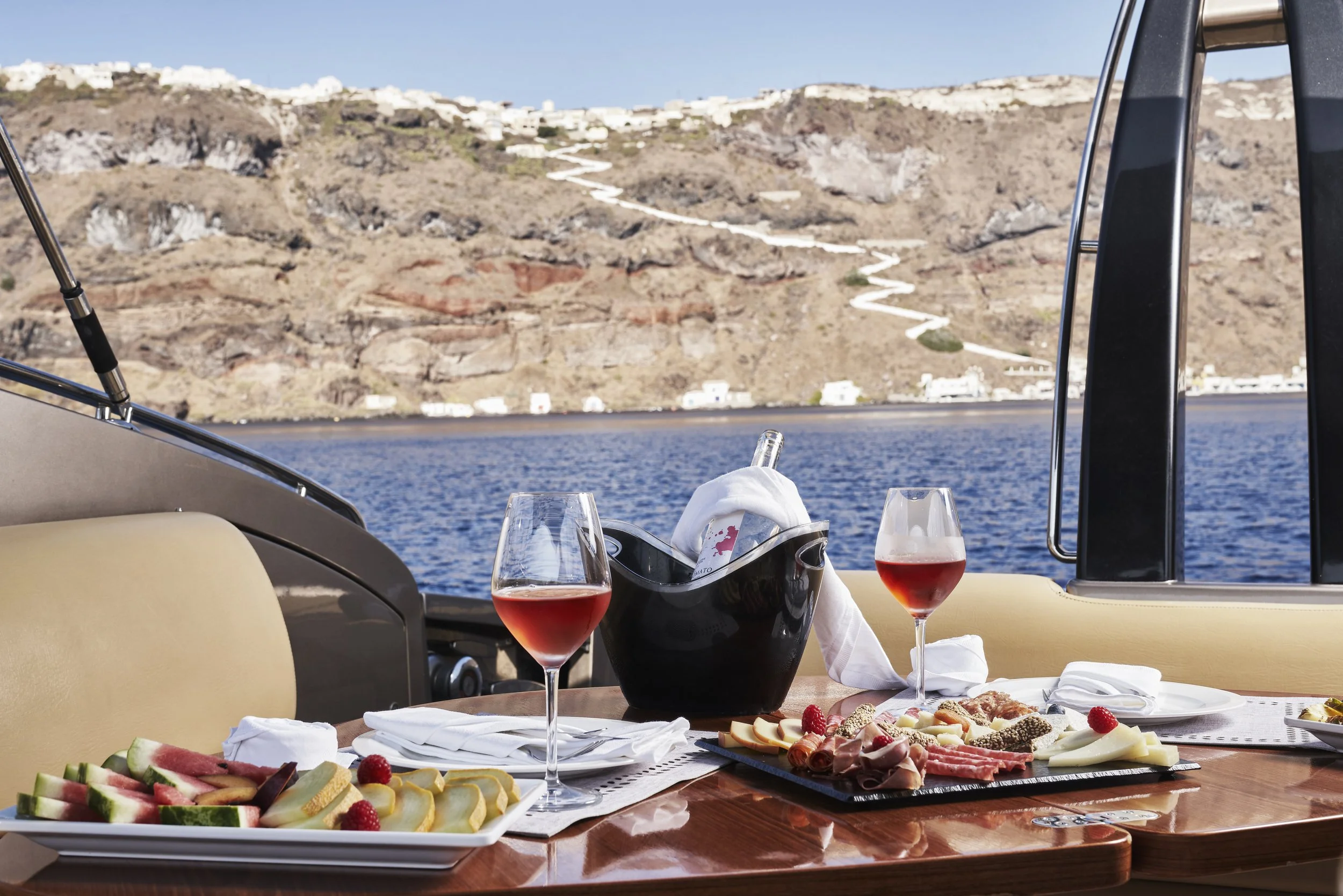 A table on a boat with two glasses of rosé wine, a black ice bucket with a bottle, and a charcuterie and cheese platter with slices of ham, salami, and cheese, set against a backdrop of a body of water and a hillside with white buildings.