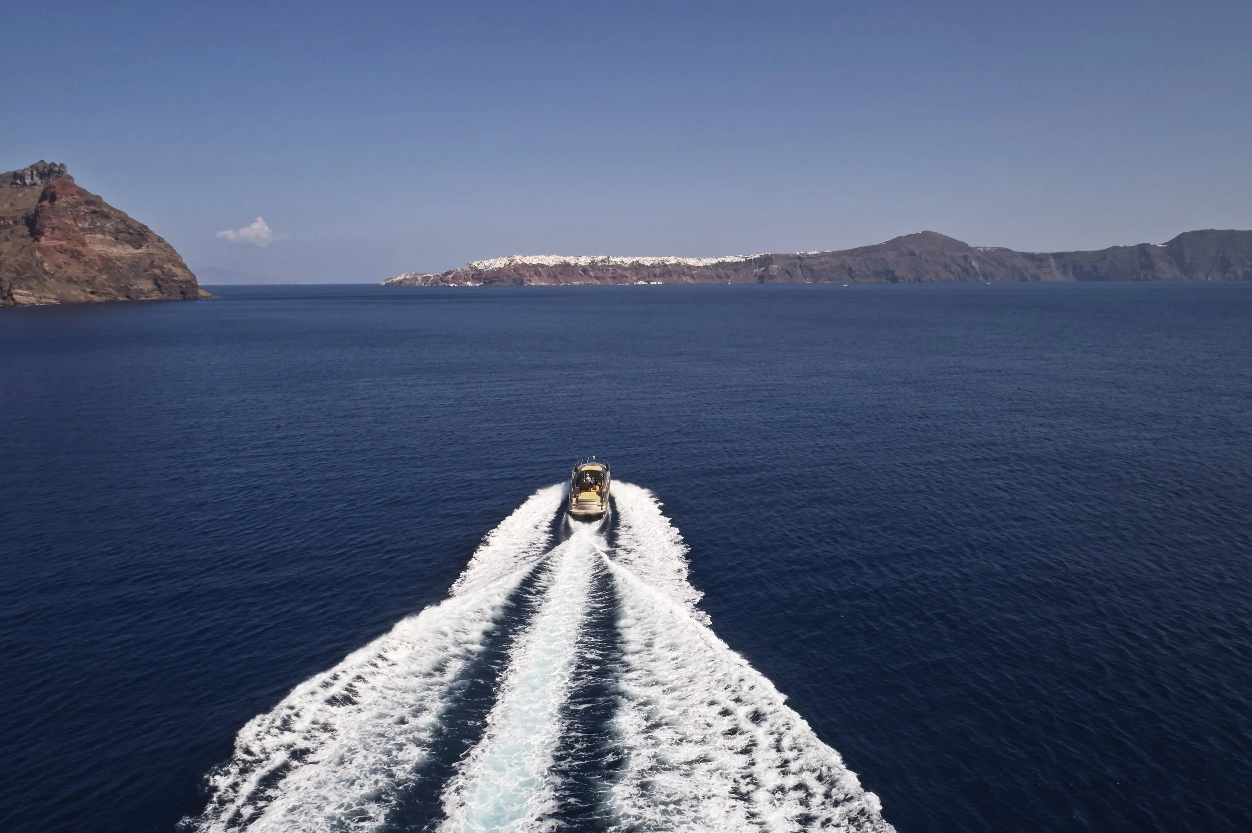 A boat speeding through deep blue sea, creating a white wake, with rocky islands in the background under a clear blue sky.