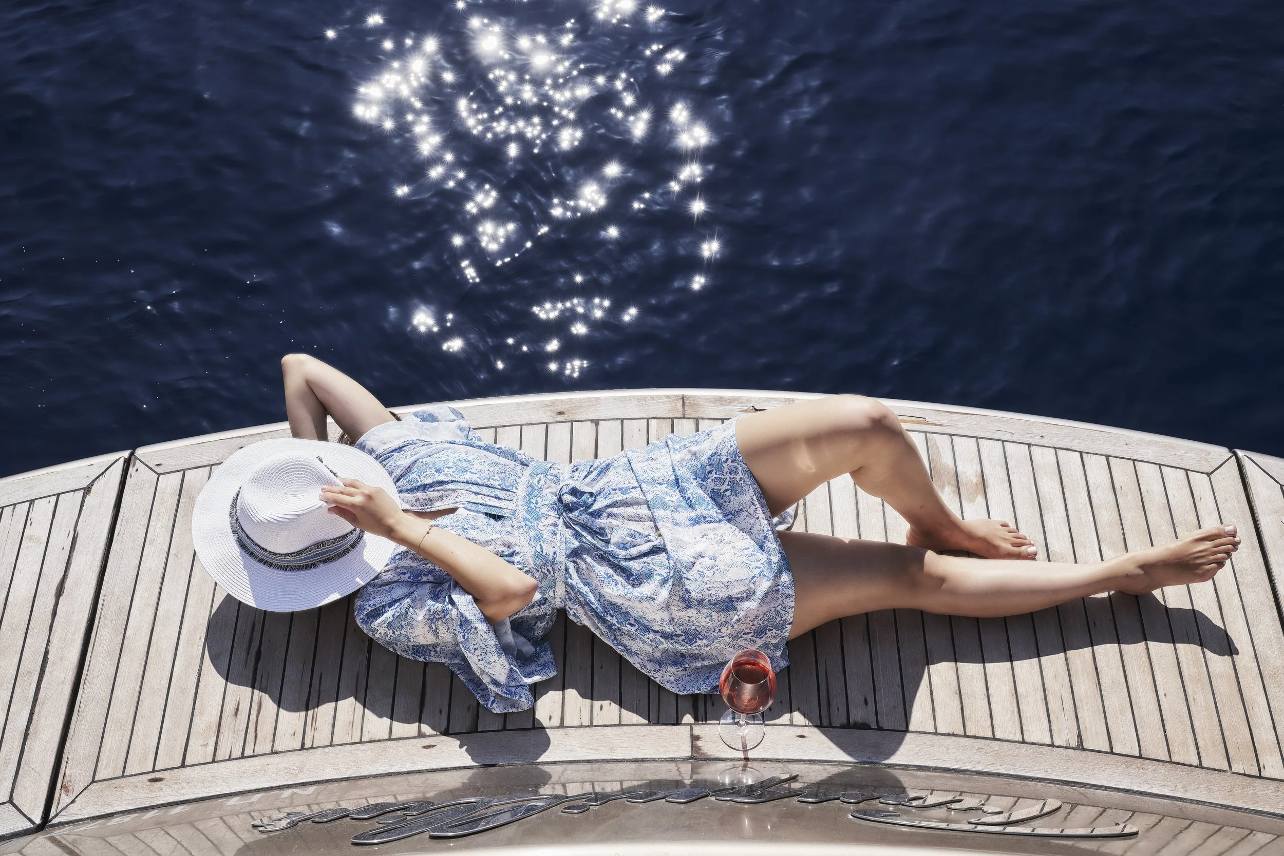 A woman relaxing on a wooden boat deck, wearing a blue dress and a wide-brimmed white hat, with a glass of rosé wine nearby, as she lounges by the water.