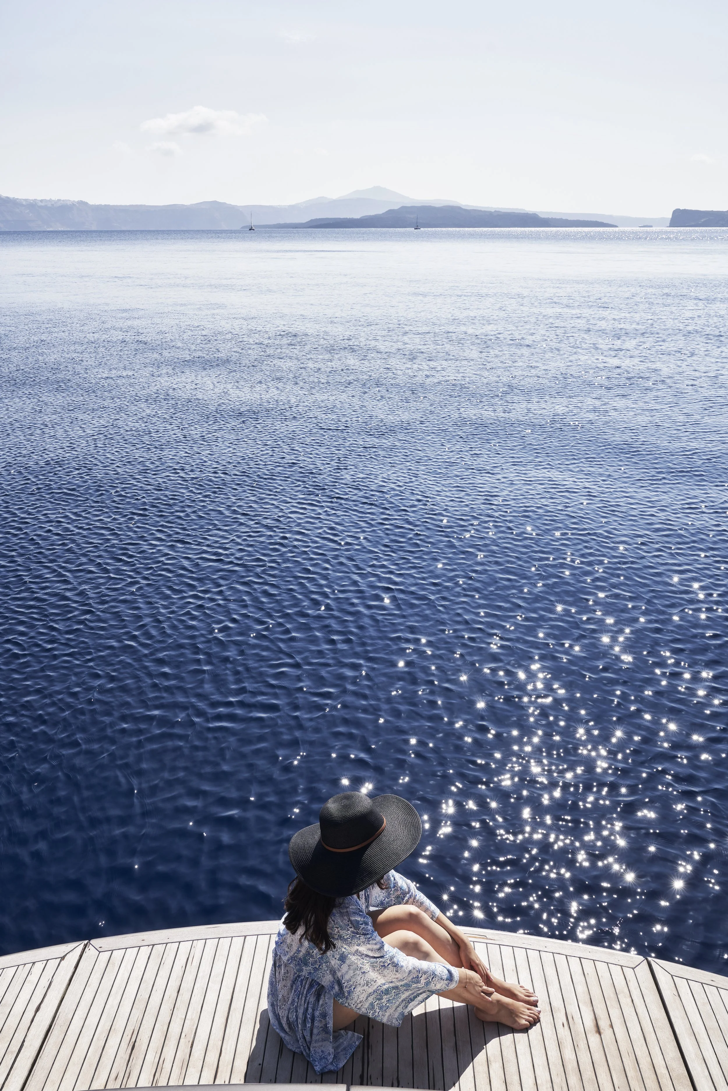 A woman with dark hair wearing a flowy blue dress and large black sunhat sits on a wooden dock by a large body of water, looking out at the calm lake with mountains and a few sailboats in the distance under a partly cloudy sky.