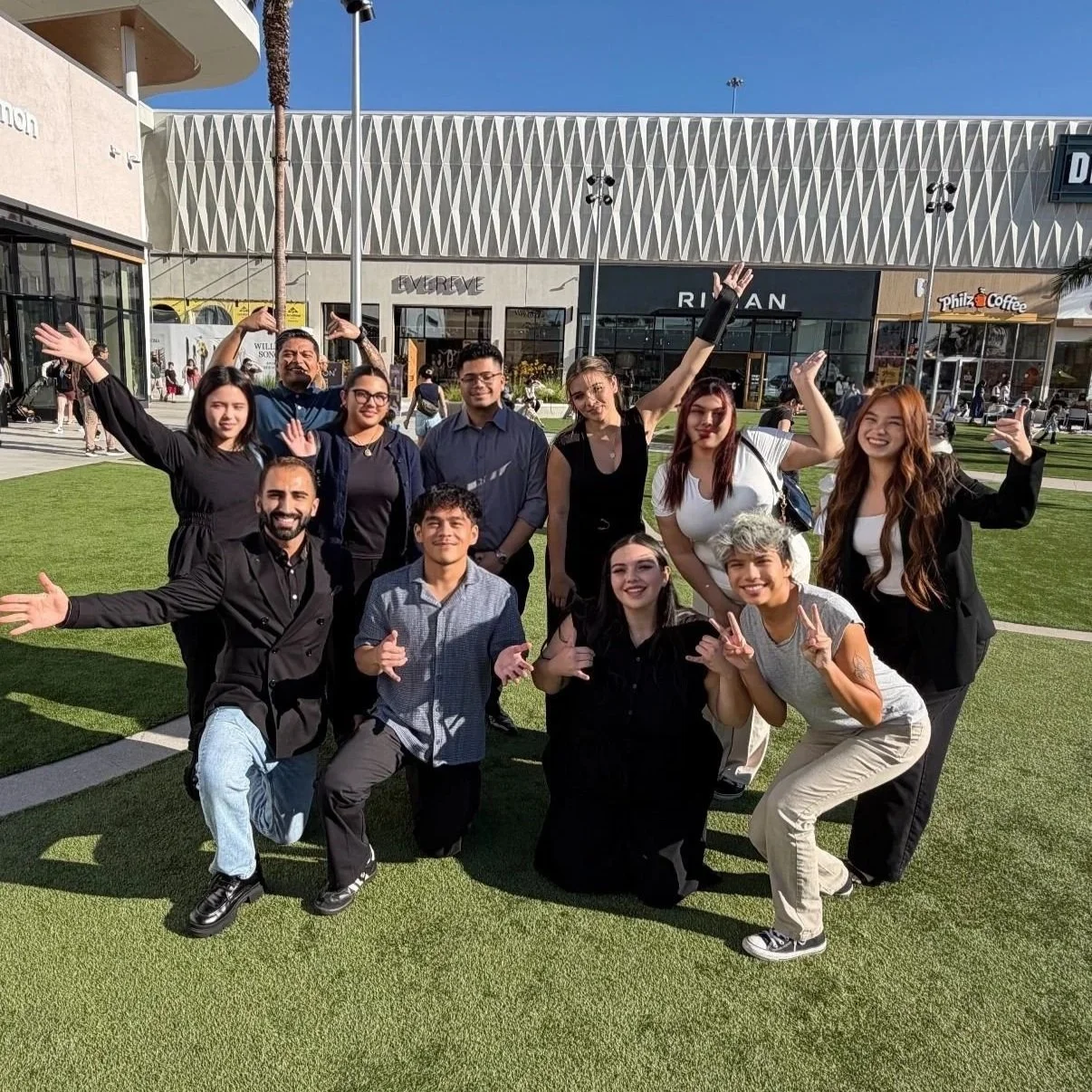A group of ten diverse young people posing outdoors on a sunny day, some crouching and some standing, making various hand gestures and smiling in front of a shopping mall.