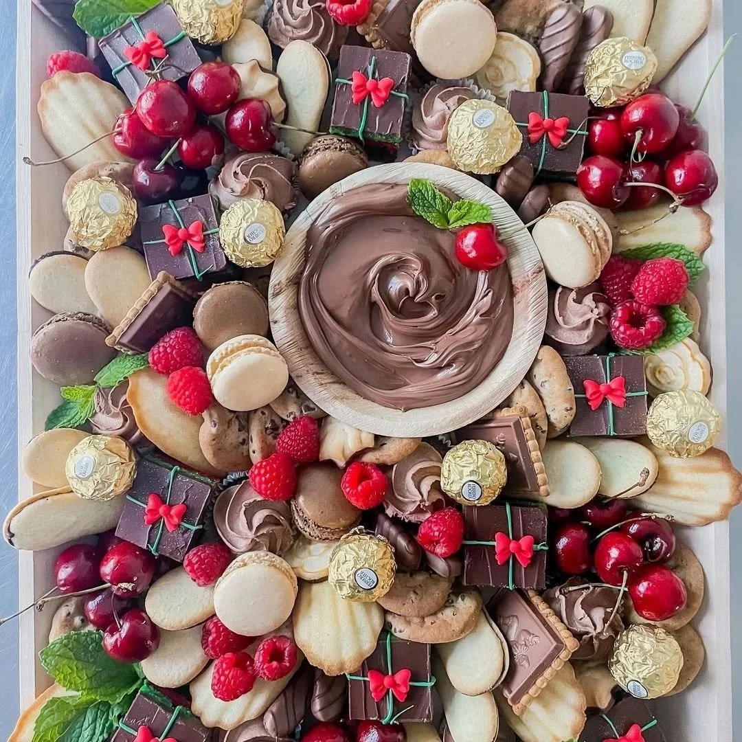 Assorted chocolates, cookies, and fresh berries arranged around a bowl of chocolate spread garnished with a cherry and mint leaves.