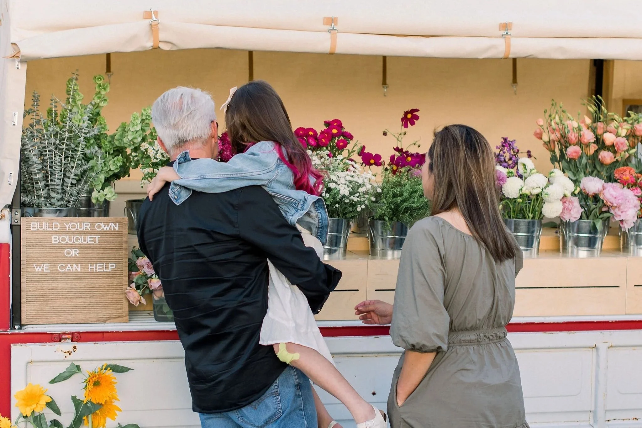 Three people at a flower stand, an older man carrying a young girl, and a woman standing nearby, with colorful flowers in buckets.