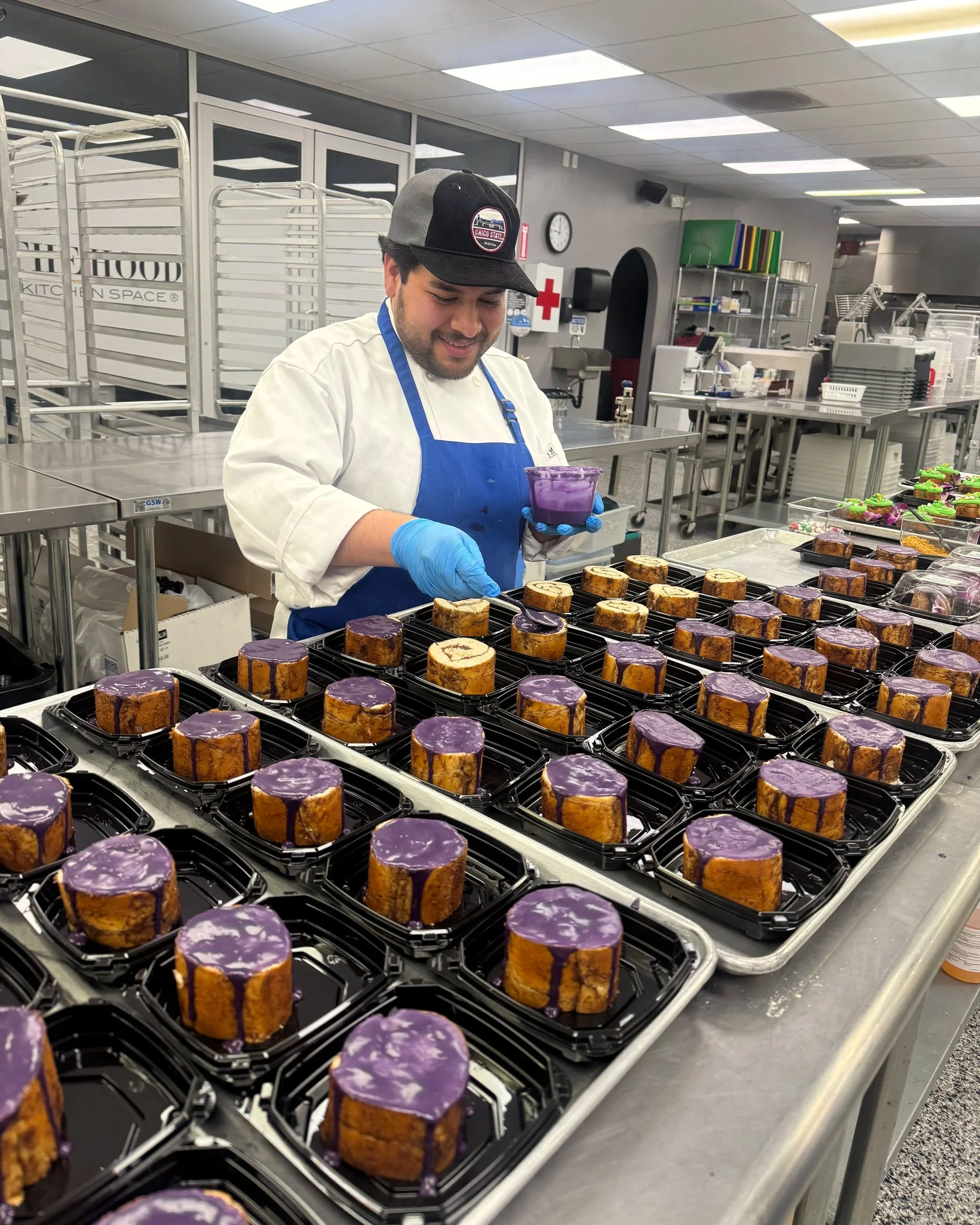 A baker in a white coat and blue apron decorating mini cakes with purple glaze in a commercial kitchen.