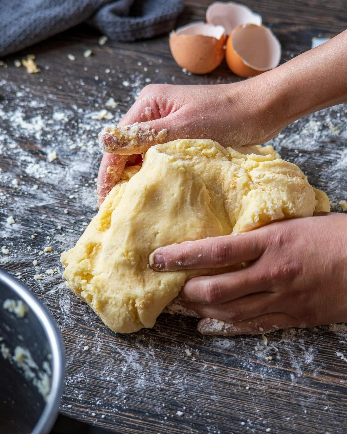 Person kneading yellow dough on a floured wooden surface with cracked eggs in the background.