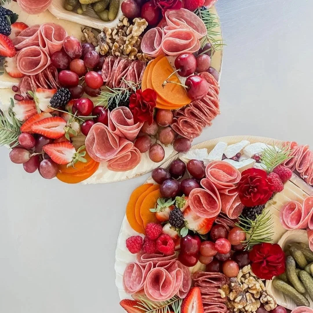 Close-up of two charcuterie boards decorated with sliced meats, cheeses, grapes, berries, strawberries, walnuts, pickles, and garnished with pine sprigs.