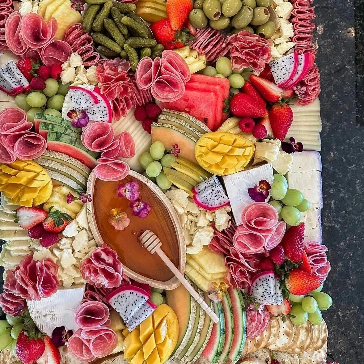 Assorted cheeses, fresh strawberries, grapes, watermelon, honey dew melon, dragon fruit, mint, and a honey dipper in a wooden honey holder.