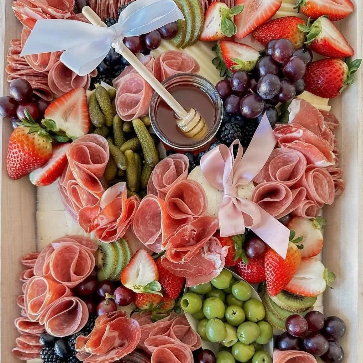 Charcuterie board with sliced strawberries, green and red grapes, honey, pickles, kiwi, blackberries, and assorted cured meats, decorated with pastel pink ribbons.