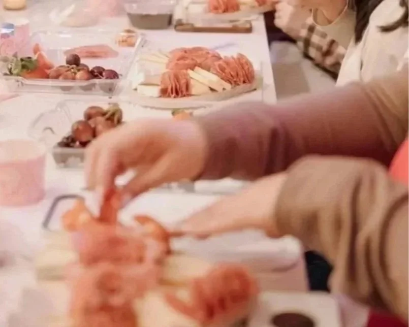 People serving themselves food at a buffet table with various dishes including sliced meats, salads, and fruits.