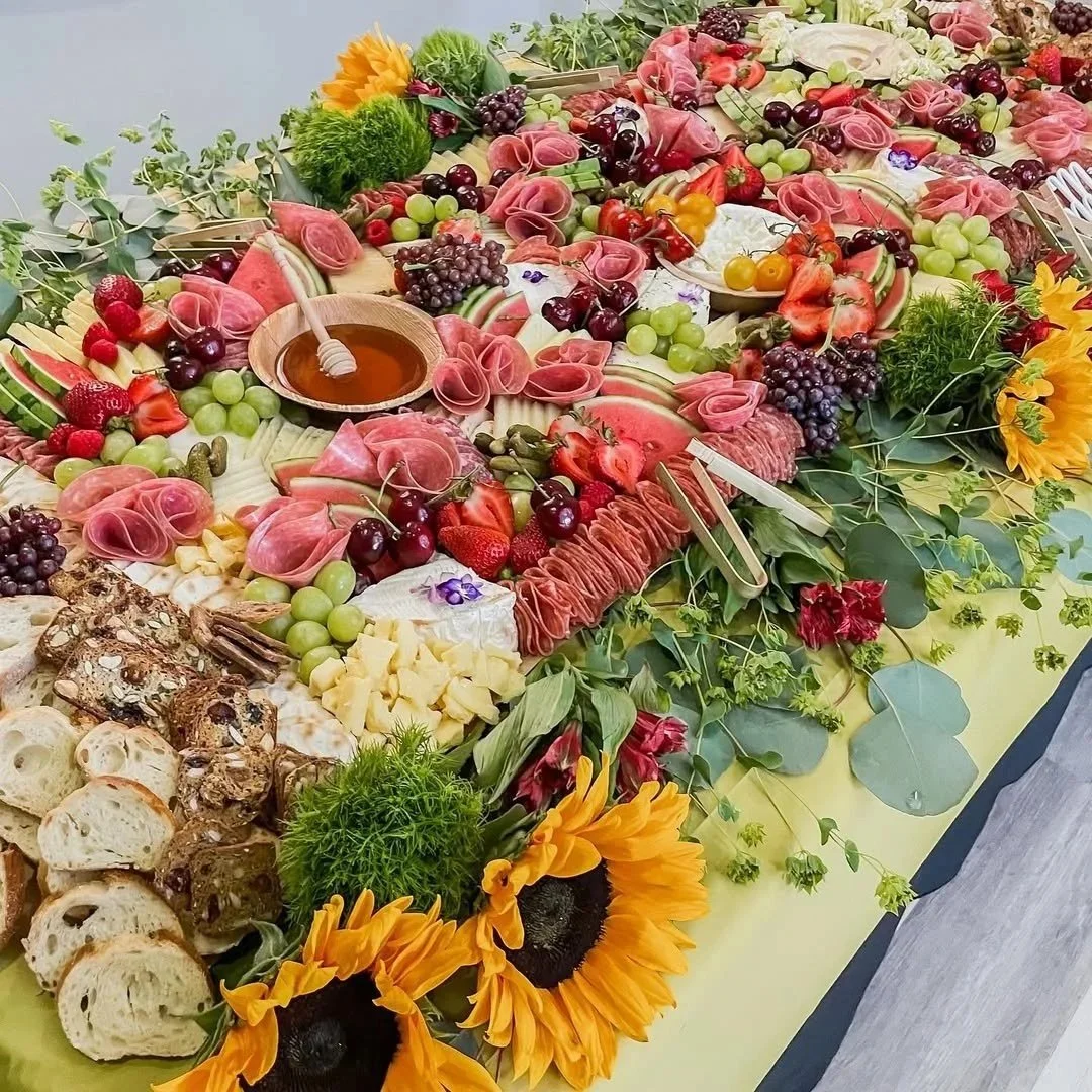 An elaborate fruit and charcuterie display featuring sliced watermelon, grapes, strawberries, cherries, various cheeses, sliced meats, bread, honey, and decorated with sunflowers, greenery, and flowers.