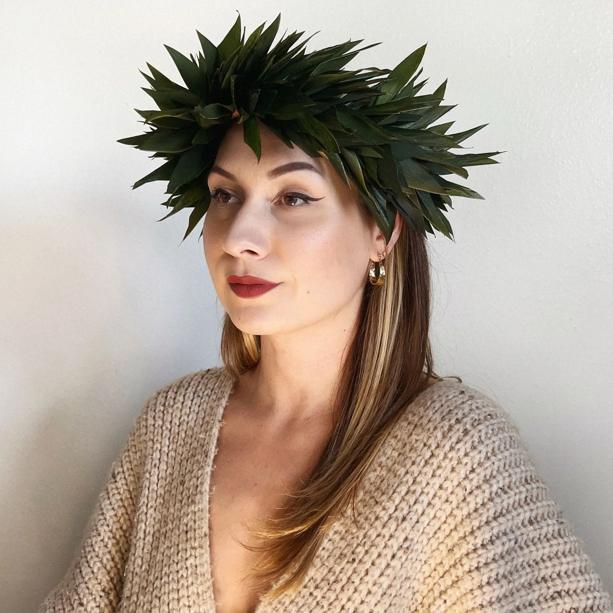 A woman wearing a crown made of green leaves, beige sweater, gold hoop earrings, and makeup with red lipstick, standing against a plain white wall.