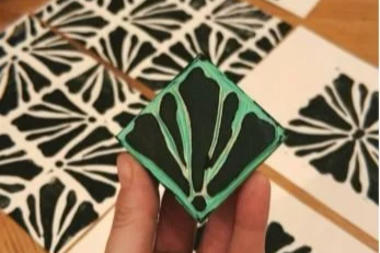 Hand holding a rubber stamp with a leaf pattern, surrounded by stamped leaf pattern prints on paper.