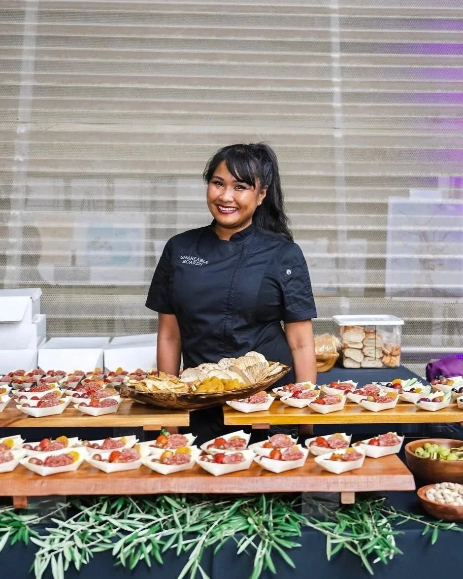 A woman in a black chef jacket standing behind a table with assorted appetizers and snacks, including small plates with meat and cherry tomatoes, bread, and grapes, with a decorative green foliage at the front.