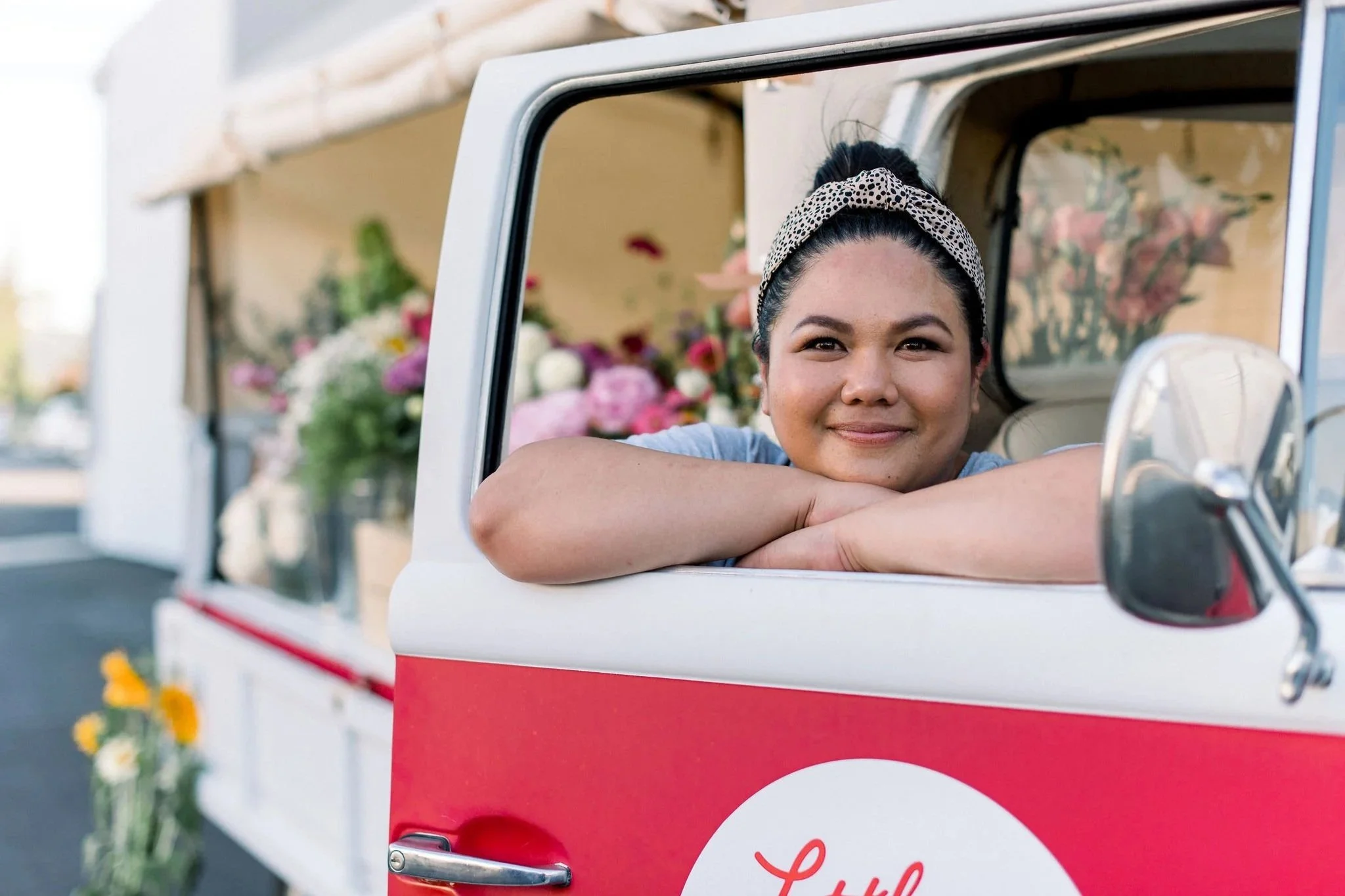 A woman with a patterned headband resting her arms on the window ledge of a red and white truck. The truck is filled with colorful flowers, and outdoor setting with sunlight and flowers on the side of the road.