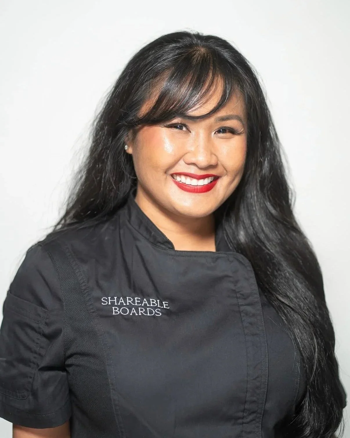 Woman with long black hair, wearing a black chef's coat with 'SHAREABLE BOARDS' embroidered on it, smiling against a white background.