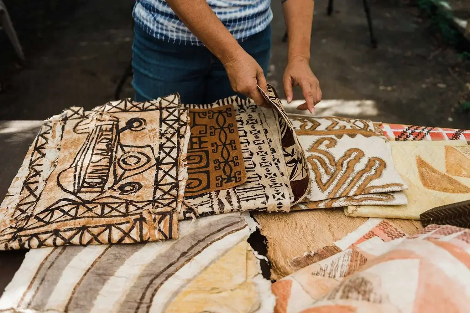 Person displaying colorful patterned textiles or traditional fabrics on a wooden table at an outdoor market.