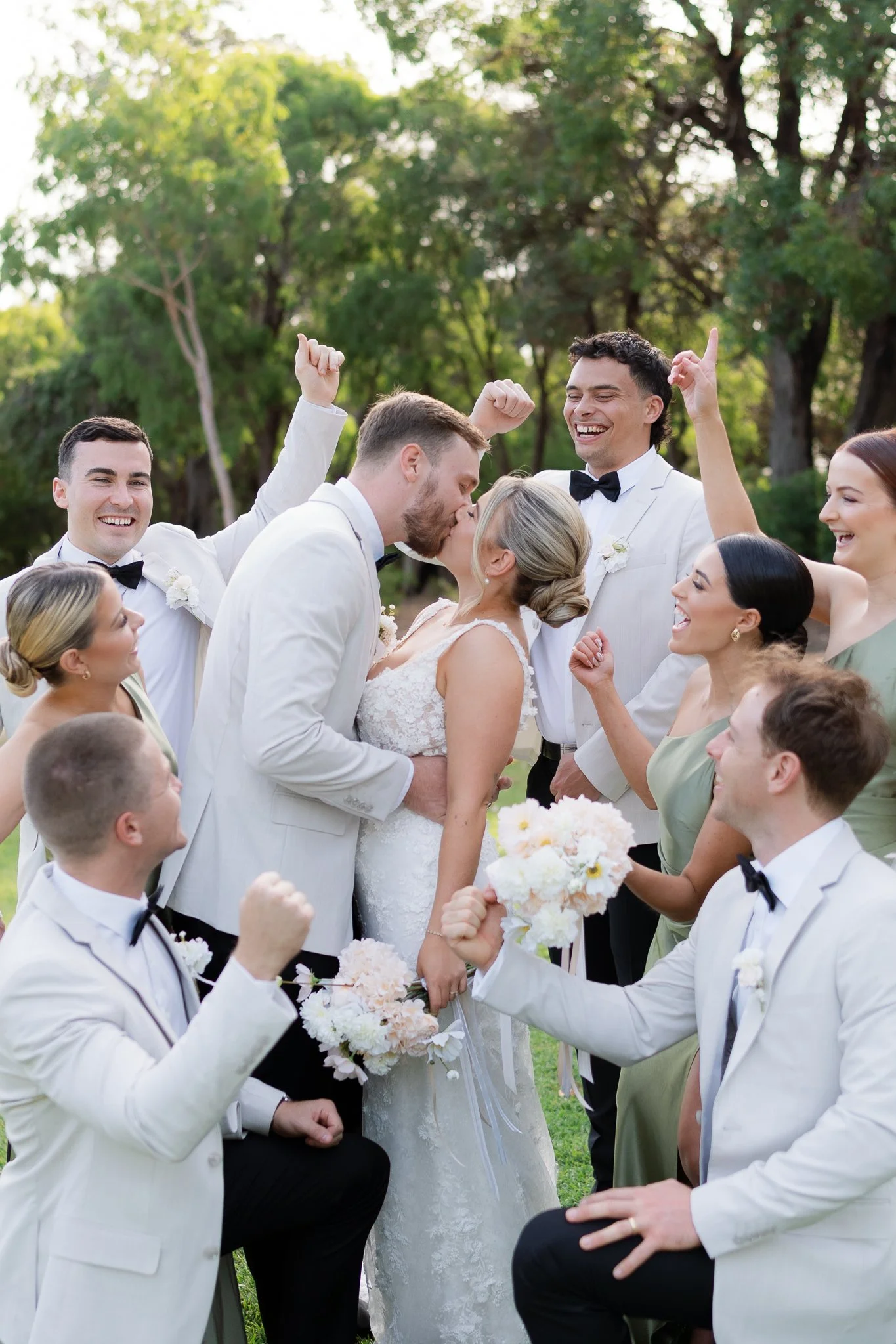 Couple kissing while surrounded by friends at a wedding celebration outdoors, with everyone smiling and cheering.