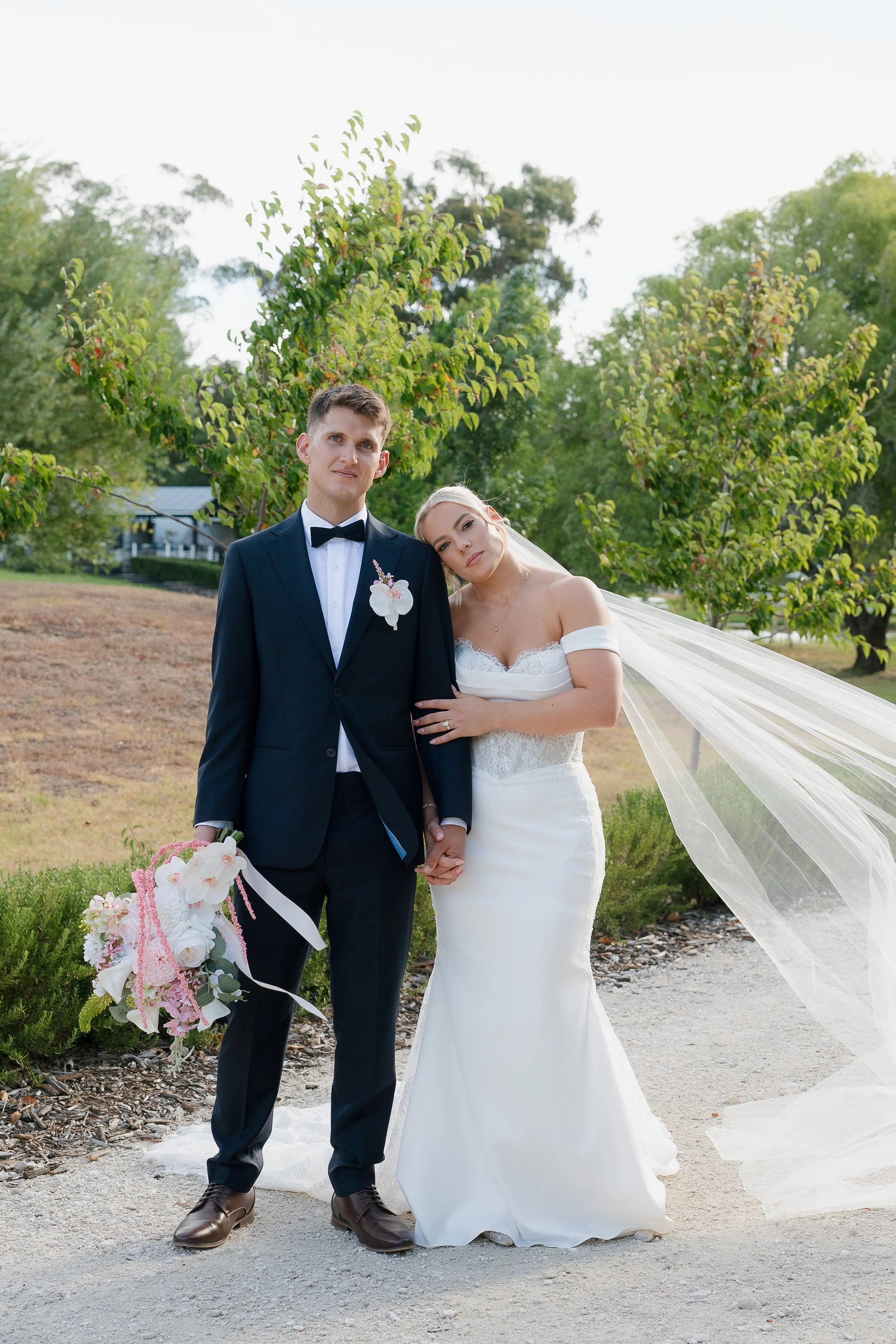 A bride and groom holding hands outdoors. The groom is dressed in a dark tuxedo with a bow tie, holding a bouquet of flowers. The bride is in a white strapless wedding gown with a veil, leaning on the groom's shoulder, in front of green trees and a garden path.