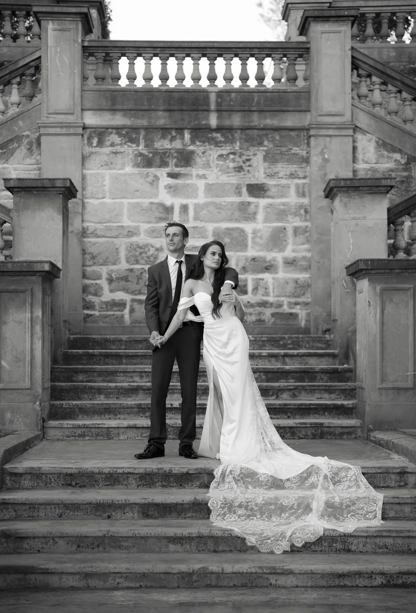 A black and white photo of a bride and groom standing on stone steps in front of an old stone building with a decorative balustrade. The groom is in a tuxedo and the bride is in a strapless wedding gown with a lace train.