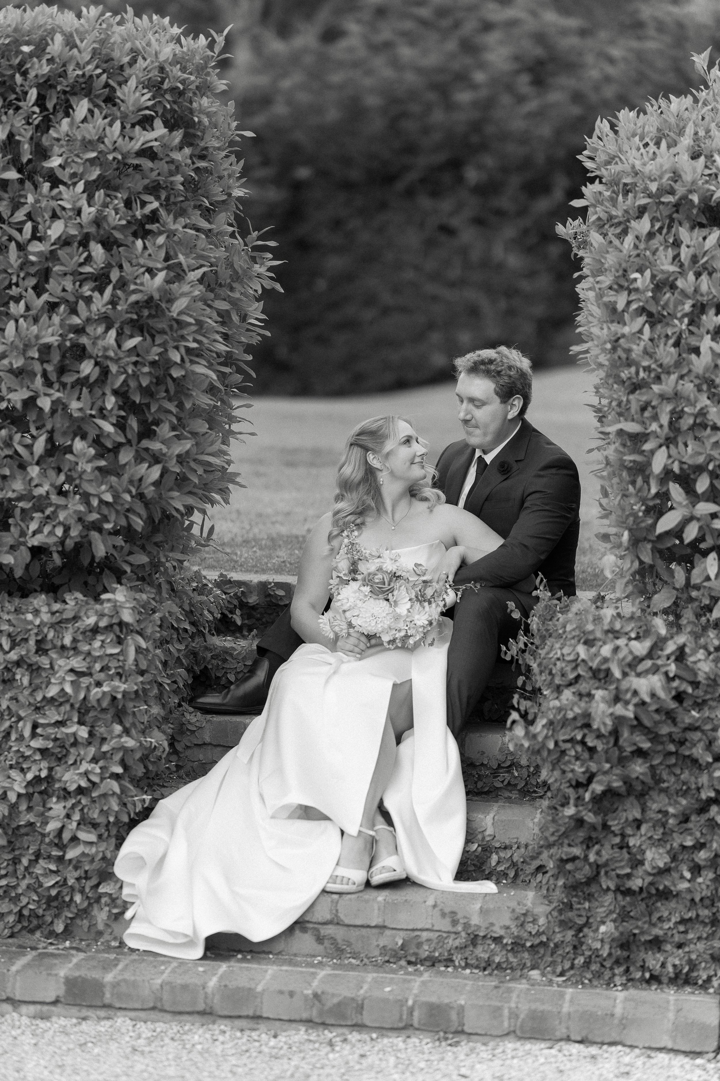 Black and white photo of a bride and groom sitting on steps, gazing into each other's eyes, surrounded by large bushes in an outdoor setting.
