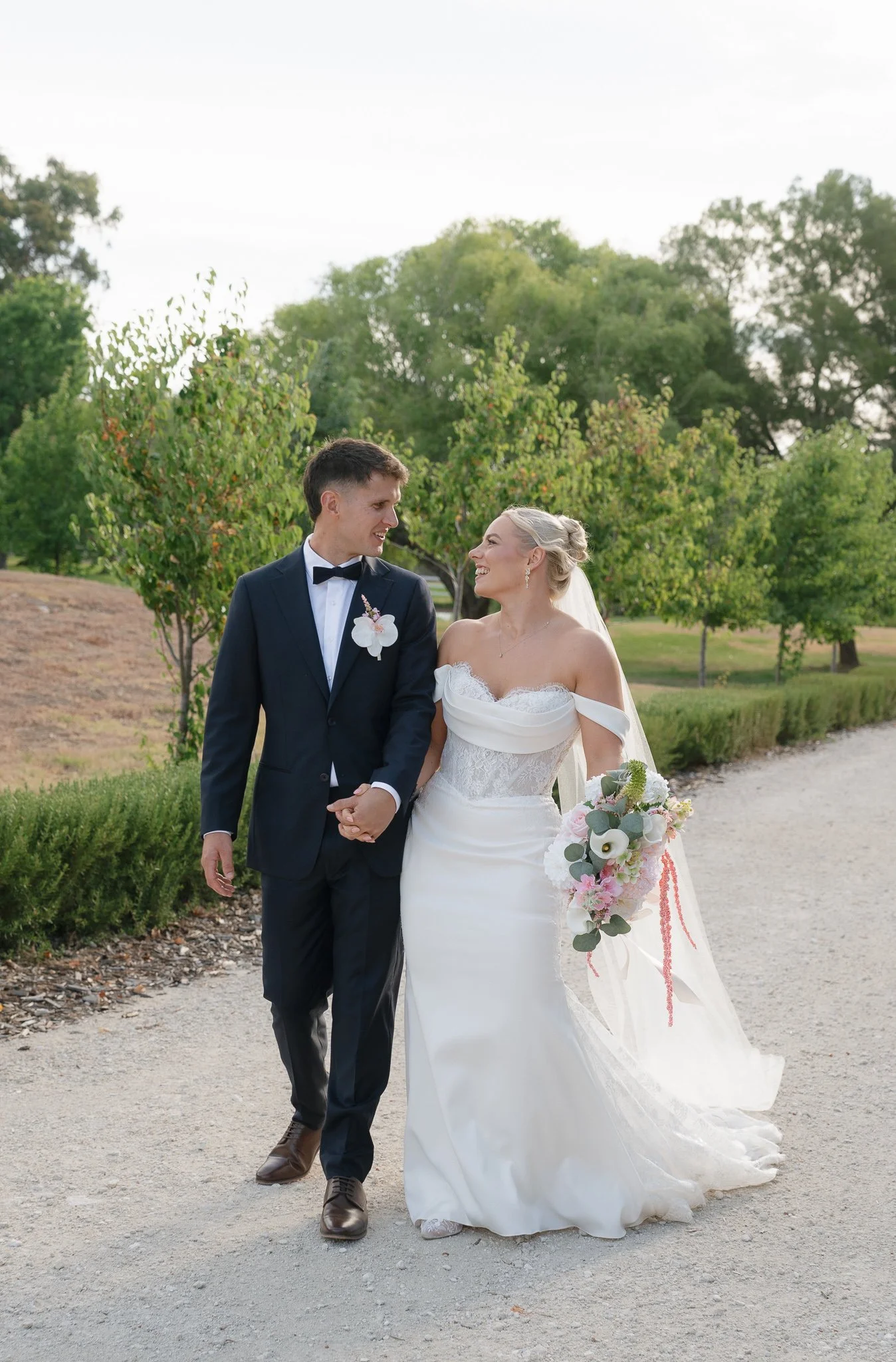 A bride and groom walking outdoors on a gravel path, holding hands, smiling, and looking at each other. The bride is wearing a white strapless wedding gown with lace details, holding a bouquet of flowers. The groom is dressed in a dark suit with a bow tie and a white boutonniere. There are green trees and bushes in the background on a bright day.
