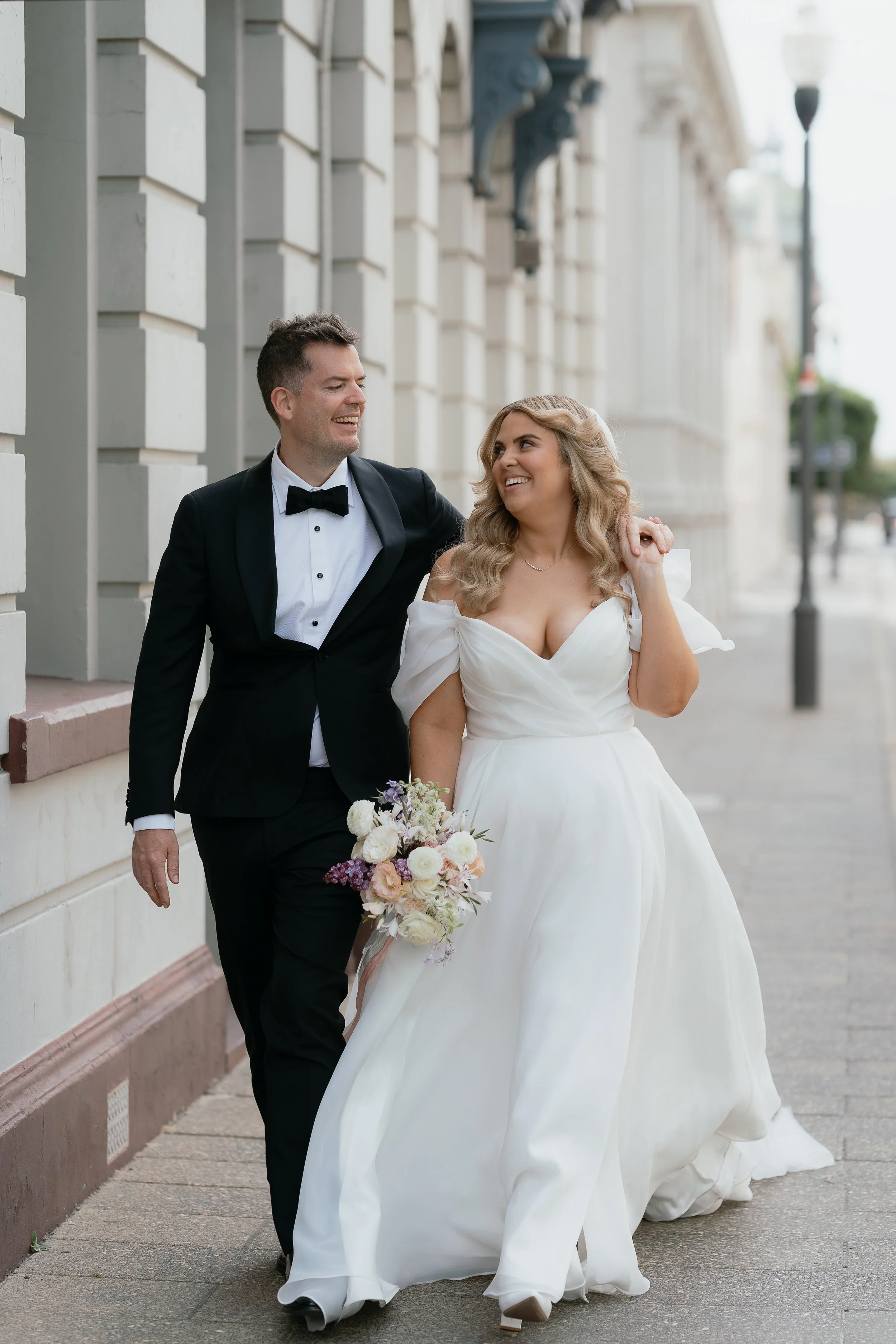 A happy bride and groom walking arm in arm down a city sidewalk, smiling at each other, with the bride holding a bouquet of flowers.