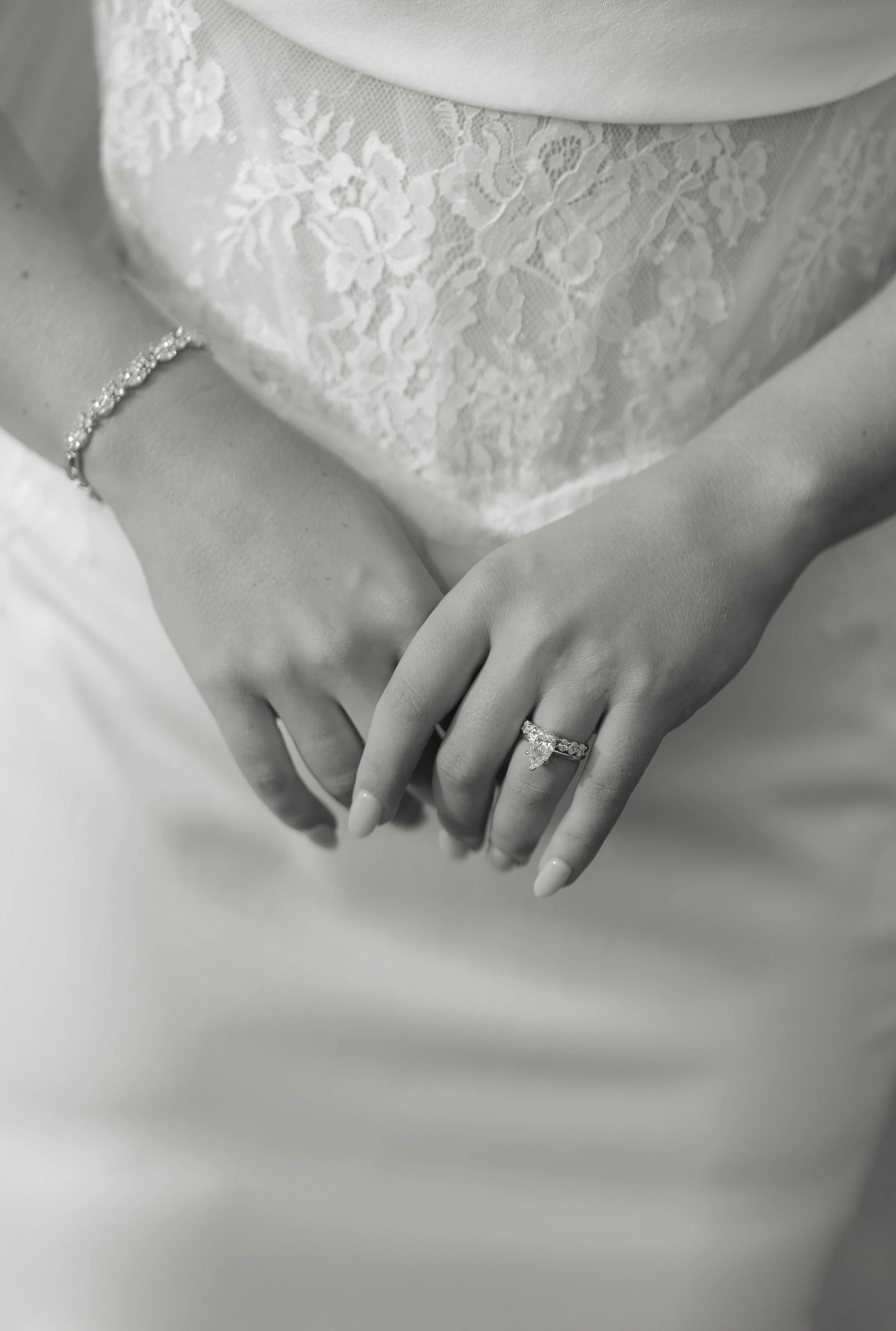 Close-up of a bride's hands with a wedding ring, bracelet, and lace dress, with the focus on her engagement ring and wedding band, in black and white.