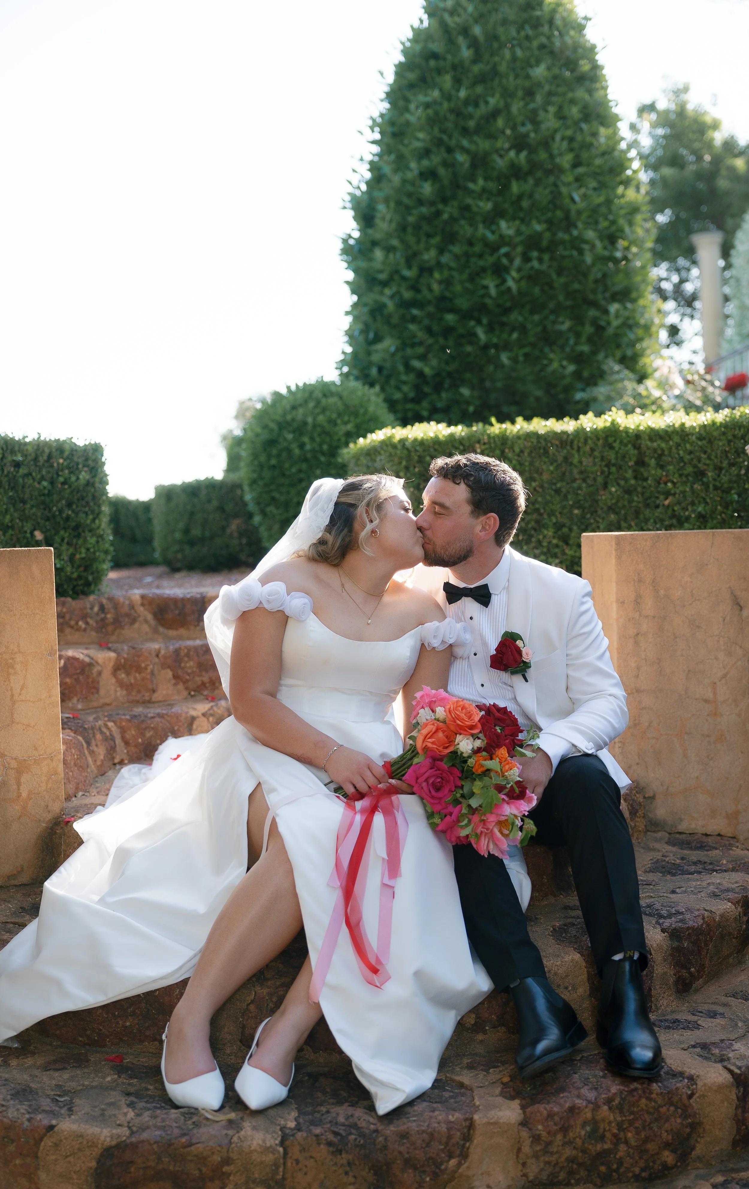 A newly married couple sharing a kiss outdoors, sitting on stone steps, with the bride holding a colorful bouquet of roses, surrounded by lush green bushes and trees.