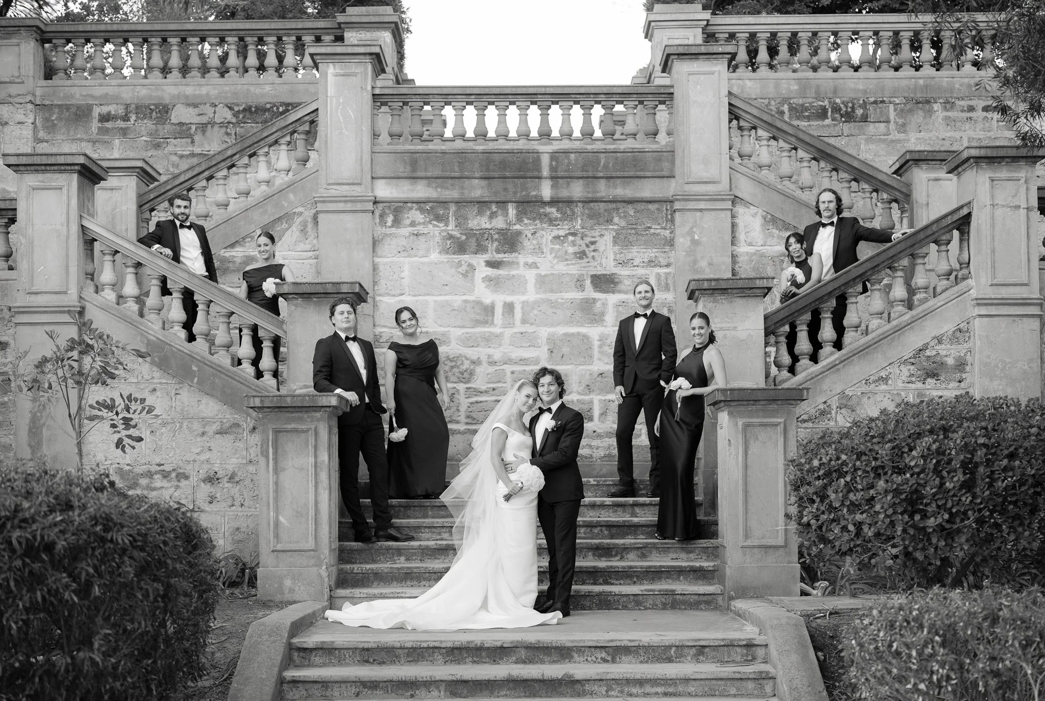 A black and white photo of a wedding party on outdoor stone staircase with ornate balustrades, with the bride and groom at the center, surrounded by bridesmaids and groomsmen in formal attire.