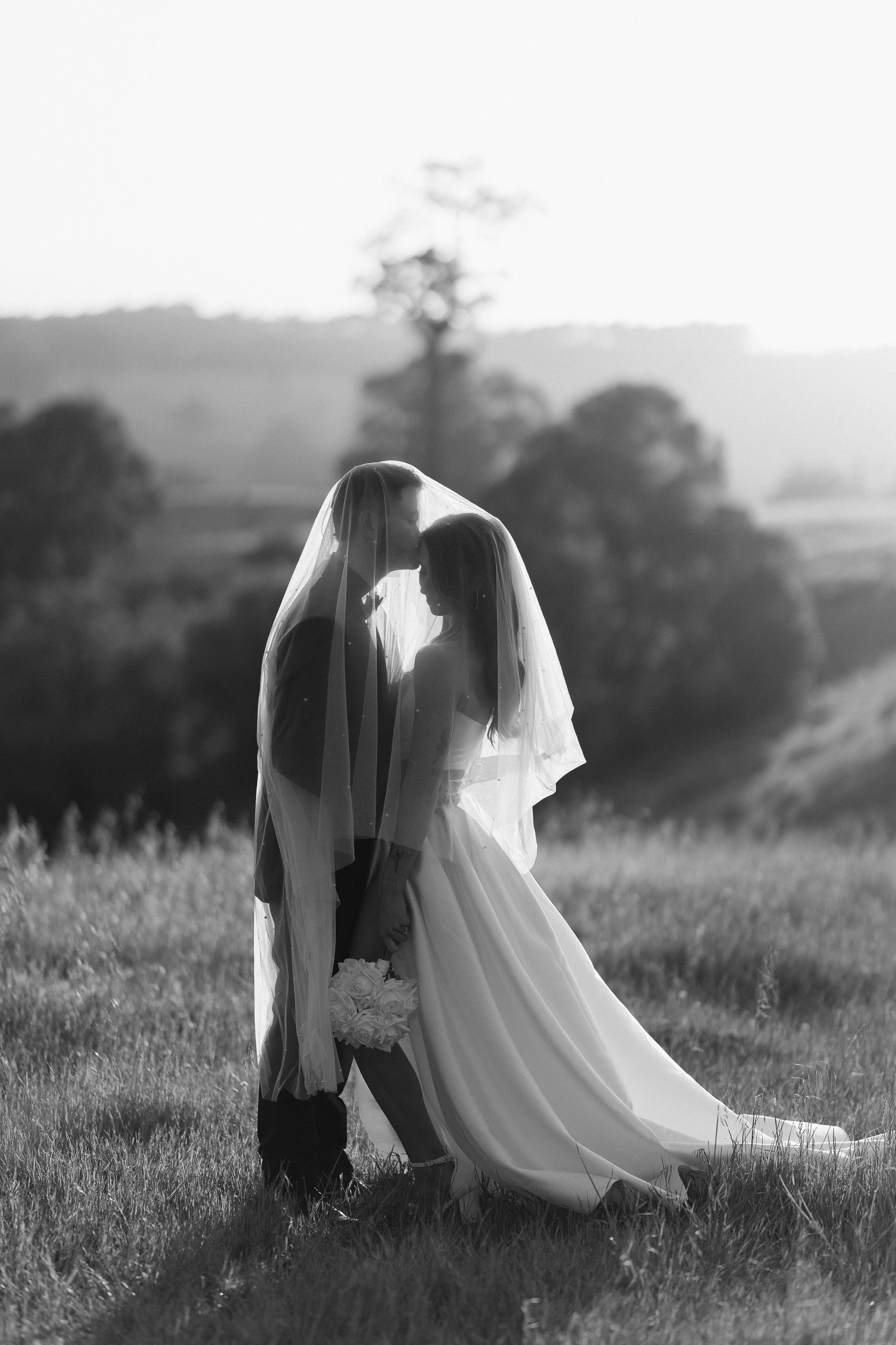 Black and white photo of a bride and groom standing close in a field, with the bride holding a bouquet of white roses. They are under a veil, touching foreheads, with a blurred landscape background.