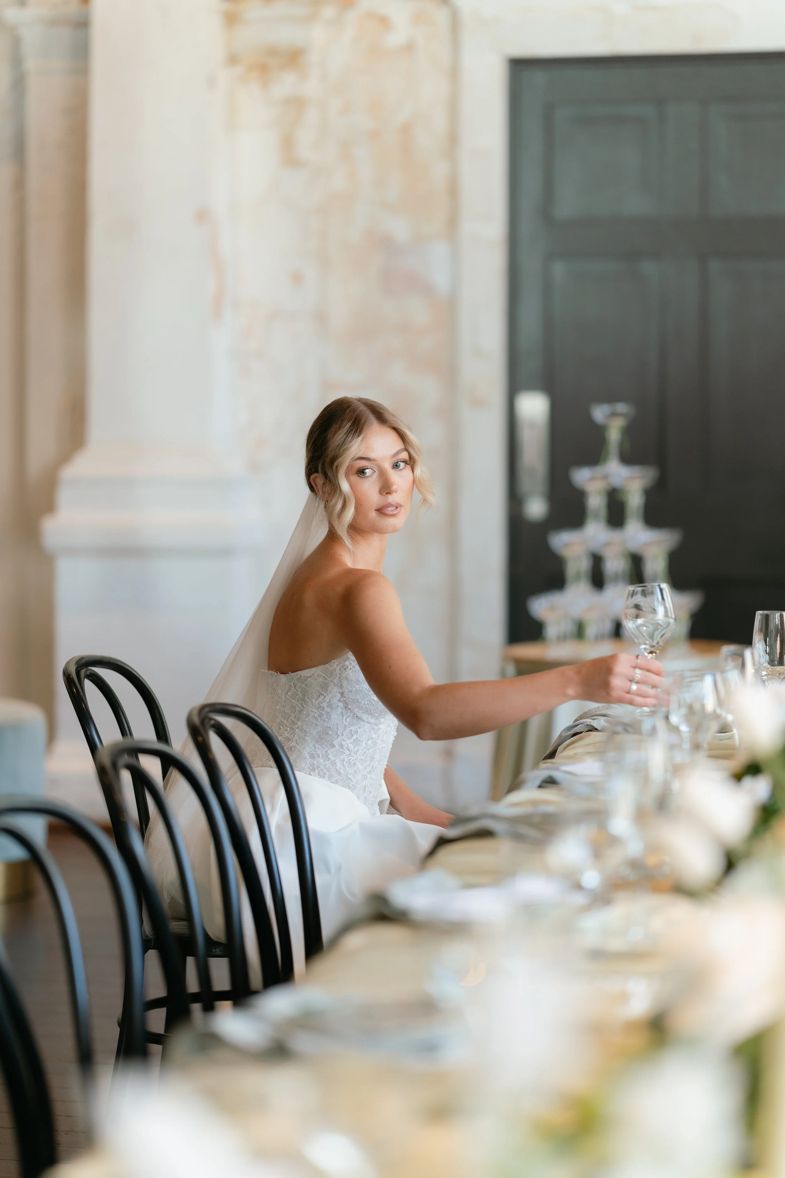 Bride in a white wedding dress sitting at a banquet table holding a wine glass, with elegant table settings and a champagne tower in the background.