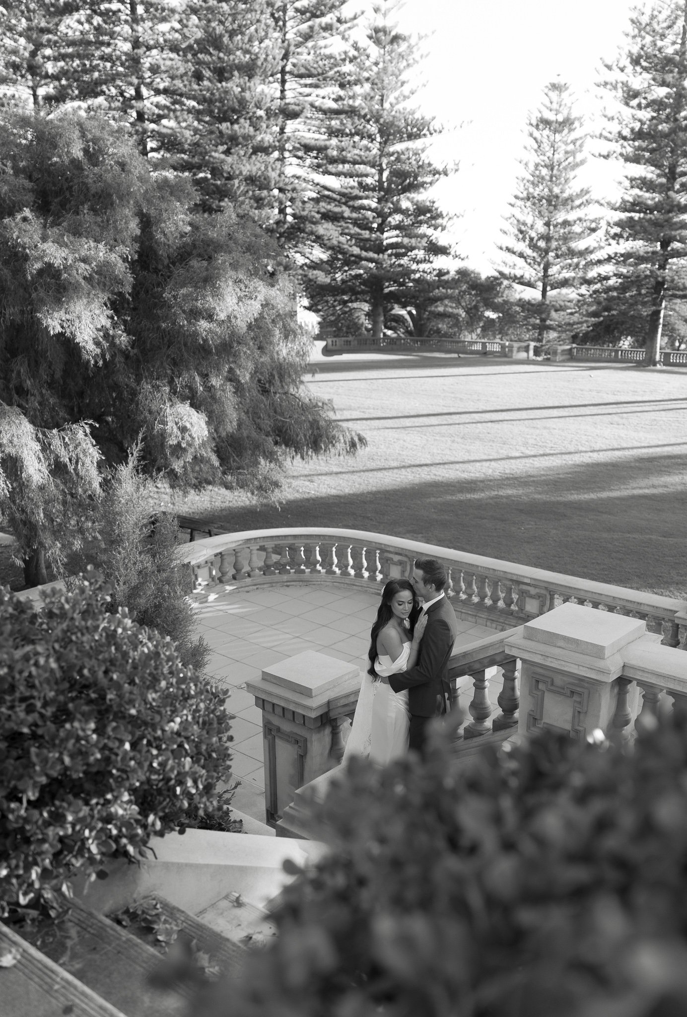 A bride and groom embrace on an elegant outdoor staircase with a decorative railing, overlooking a landscaped garden and tall pine trees in the background.