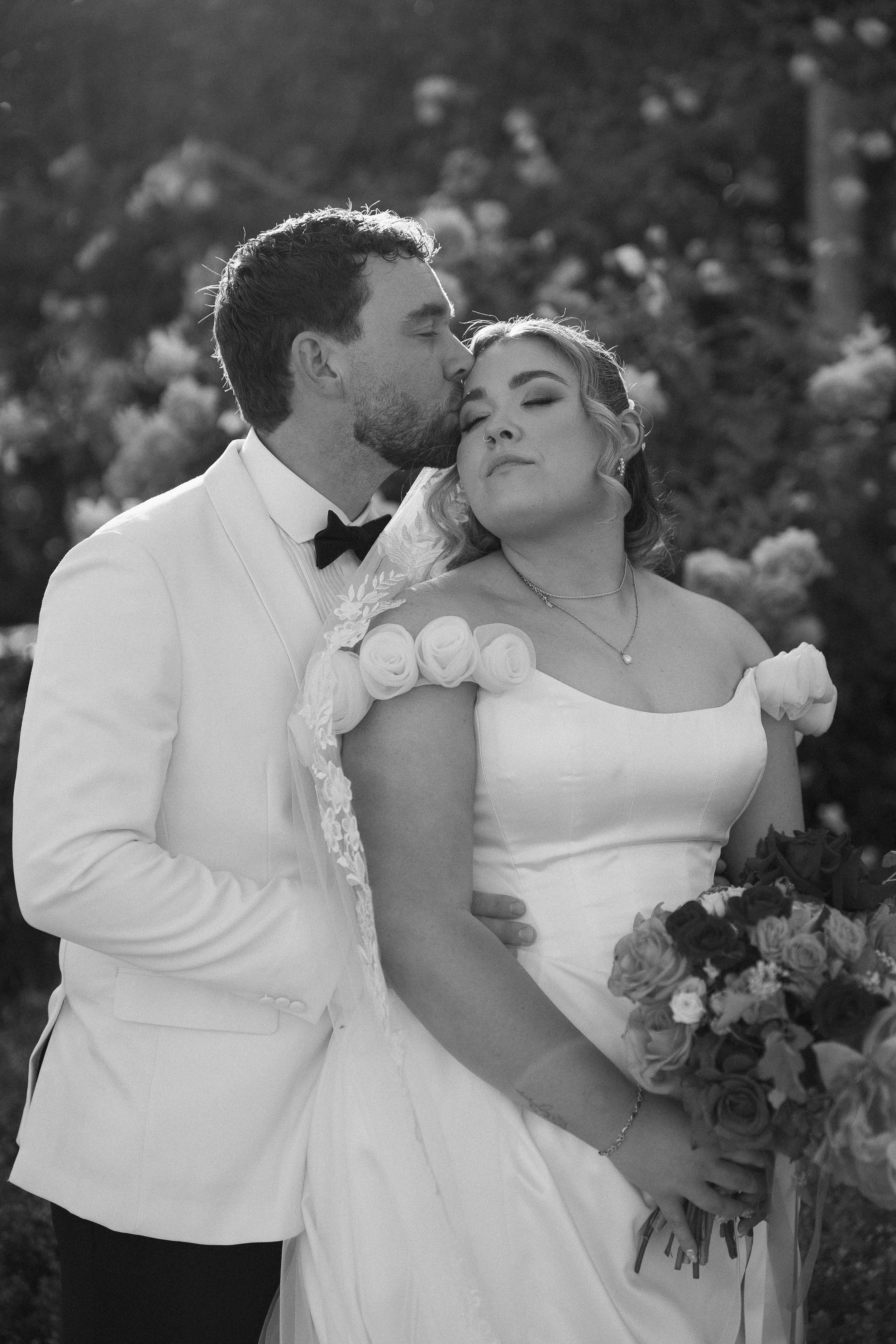 A black and white photo of a bride and groom, with the groom kissing the bride on her forehead. The bride is holding a bouquet of flowers and has her eyes closed.