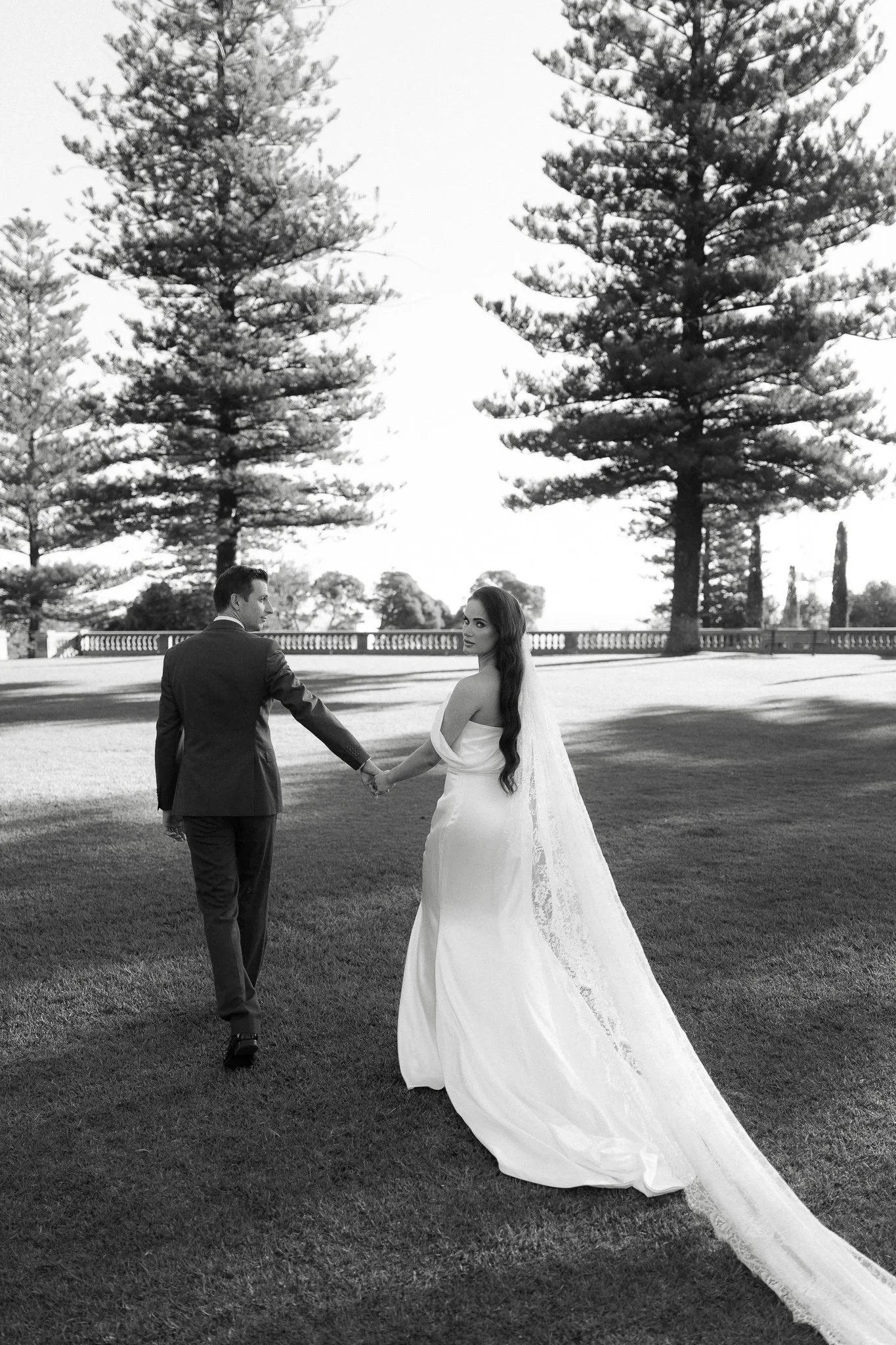 A black-and-white photograph of a couple in formal wedding attire holding hands outdoors on a grassy field, with large trees and a clear sky in the background.